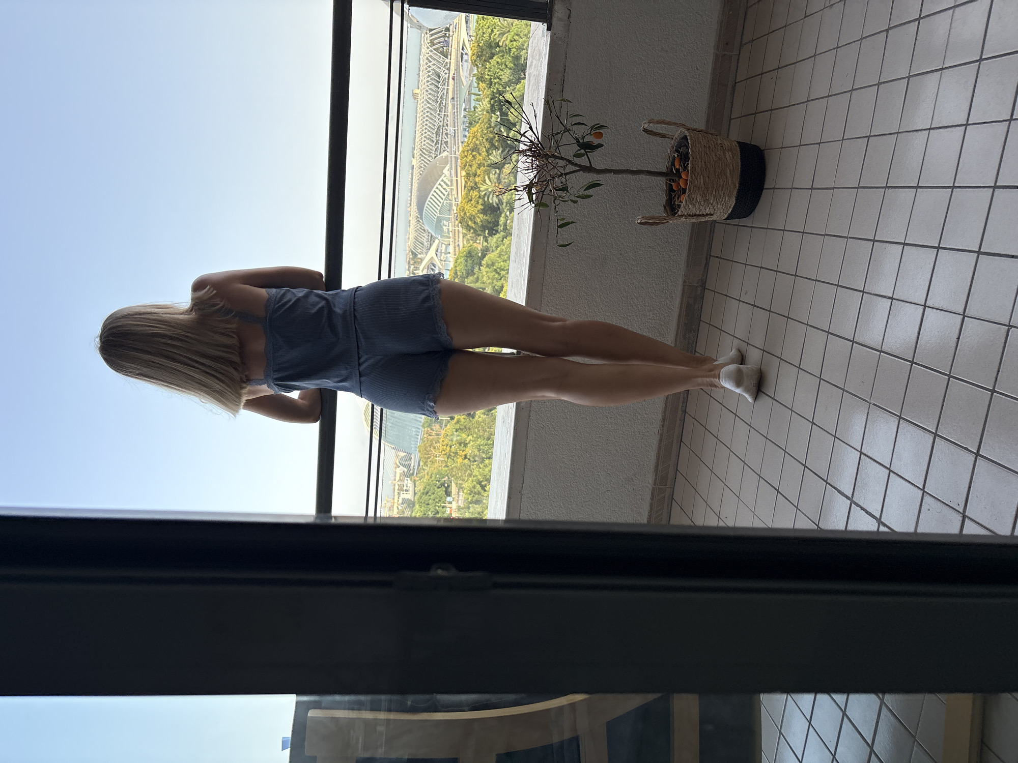 A woman stands with her back to the camera on a tiled balcony, looking out at a city view. She has blonde hair and is wearing a blue-striped romper. Her legs are long and toned, and she is wearing white socks. To her right, on the balcony, is a small potted tree with orange fruits and a woven basket. The background shows a modern cityscape with some greenery, a body of water, and a distinctive white, curved building. The sky is clear and light blue, suggesting it is daytime. The scene appears to be in València, Spain, given the architectural cues. The woman's posture suggests she is observing the view.