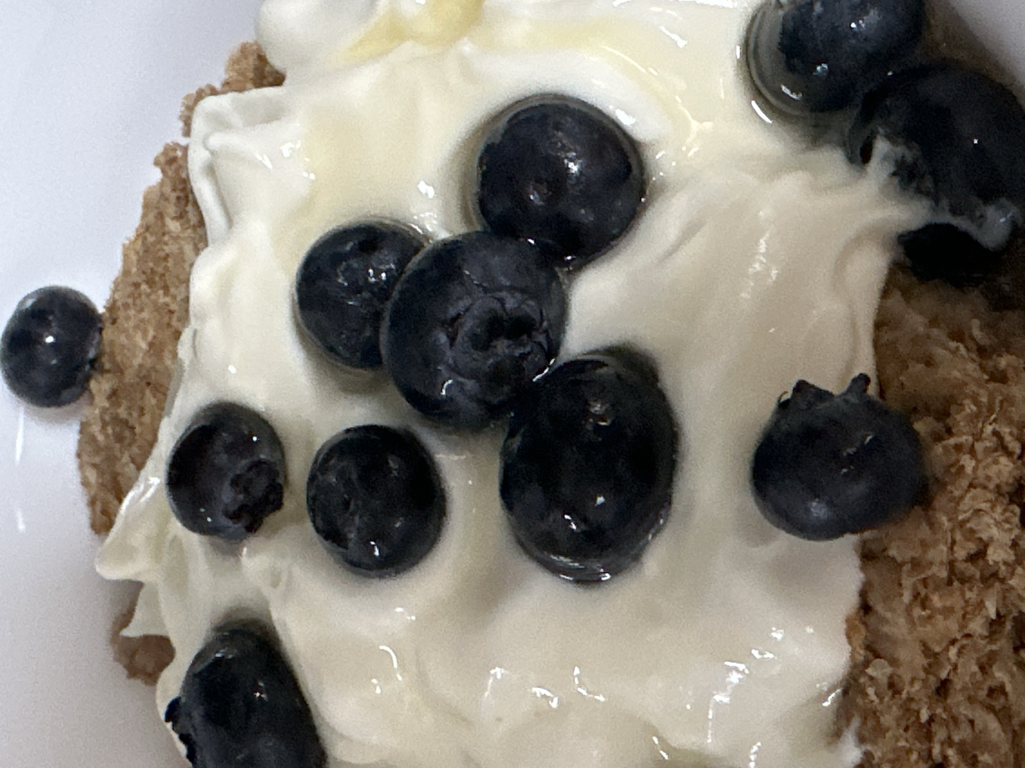 The image shows a close-up of a bowl of cereal topped with yogurt and blueberries. The cereal appears to be whole grain. The yogurt is thick and creamy, with a slightly glossy surface. Several blueberries are scattered across the yogurt, some partially submerged. The lighting is soft, highlighting the textures of the food. There are no people or any indication of the location in Moortown, Ireland.