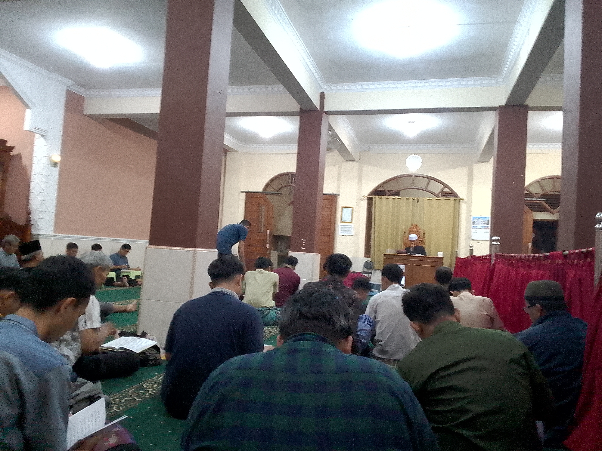 The image depicts the interior of a mosque in Kertajaya, Indonesia. Several men are seated on a green carpeted floor, some of whom are engaged in prayer or reading from religious texts. In the background, a man in a blue shirt is standing and appears to be gesturing towards something. Further back, a man in a white robe and cap is seated behind a wooden pulpit. The mosque features large, brown, square pillars and a white, decorative ceiling with overhead lighting. Red curtains are visible on the right side of the image. The overall atmosphere suggests a religious gathering or prayer service.
