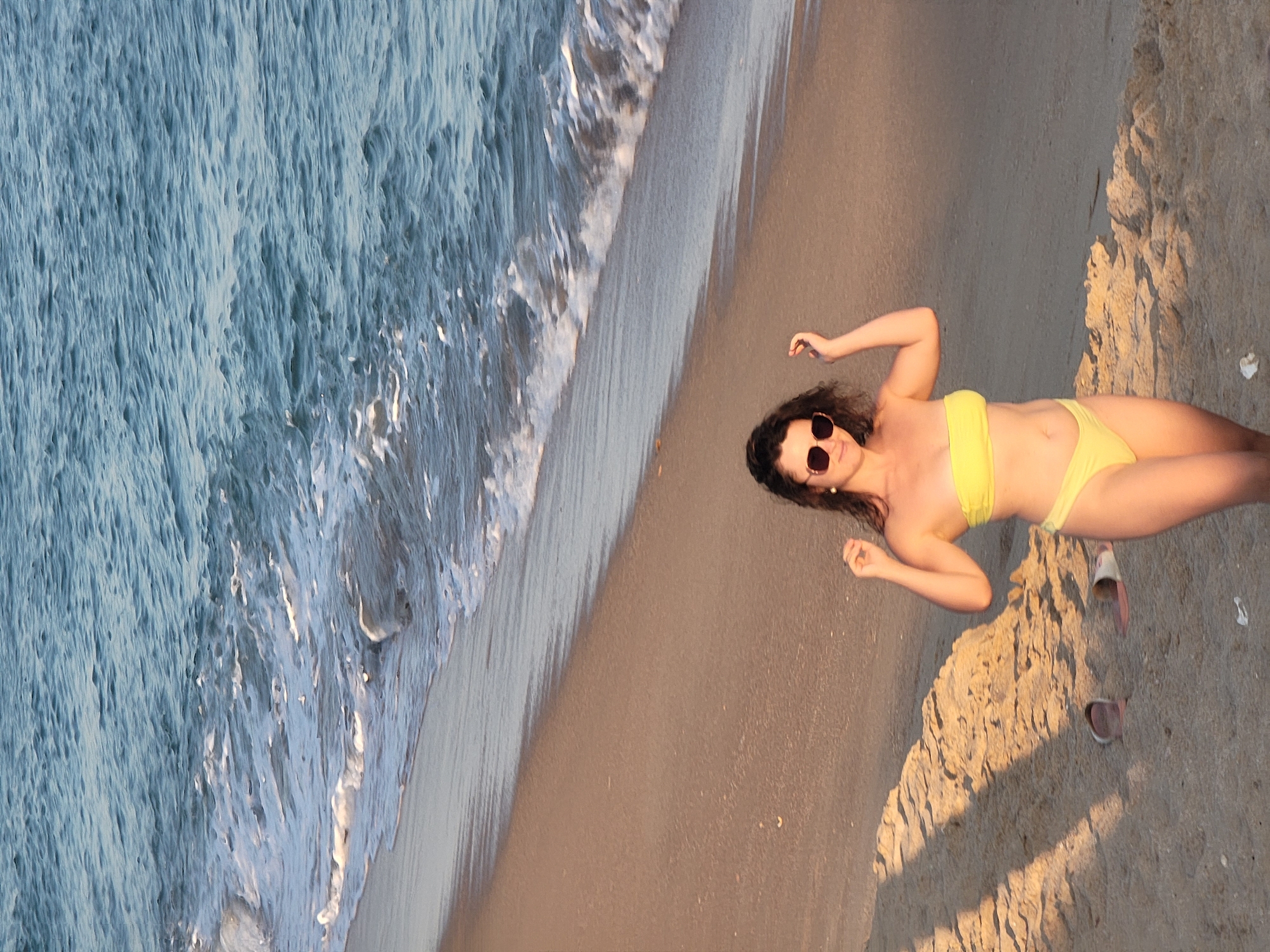 A young woman with dark, wavy hair and sunglasses stands on a sandy beach. She is wearing a yellow bikini and has her arms raised, with her hands near her head. The ocean is to her right, with gentle waves washing onto the shore. The sand has shadows cast across it, suggesting the sun is low in the sky. A pair of flip-flops are visible in the sand to her left. The scene appears to be Quarteira, Portugal, given the context provided.