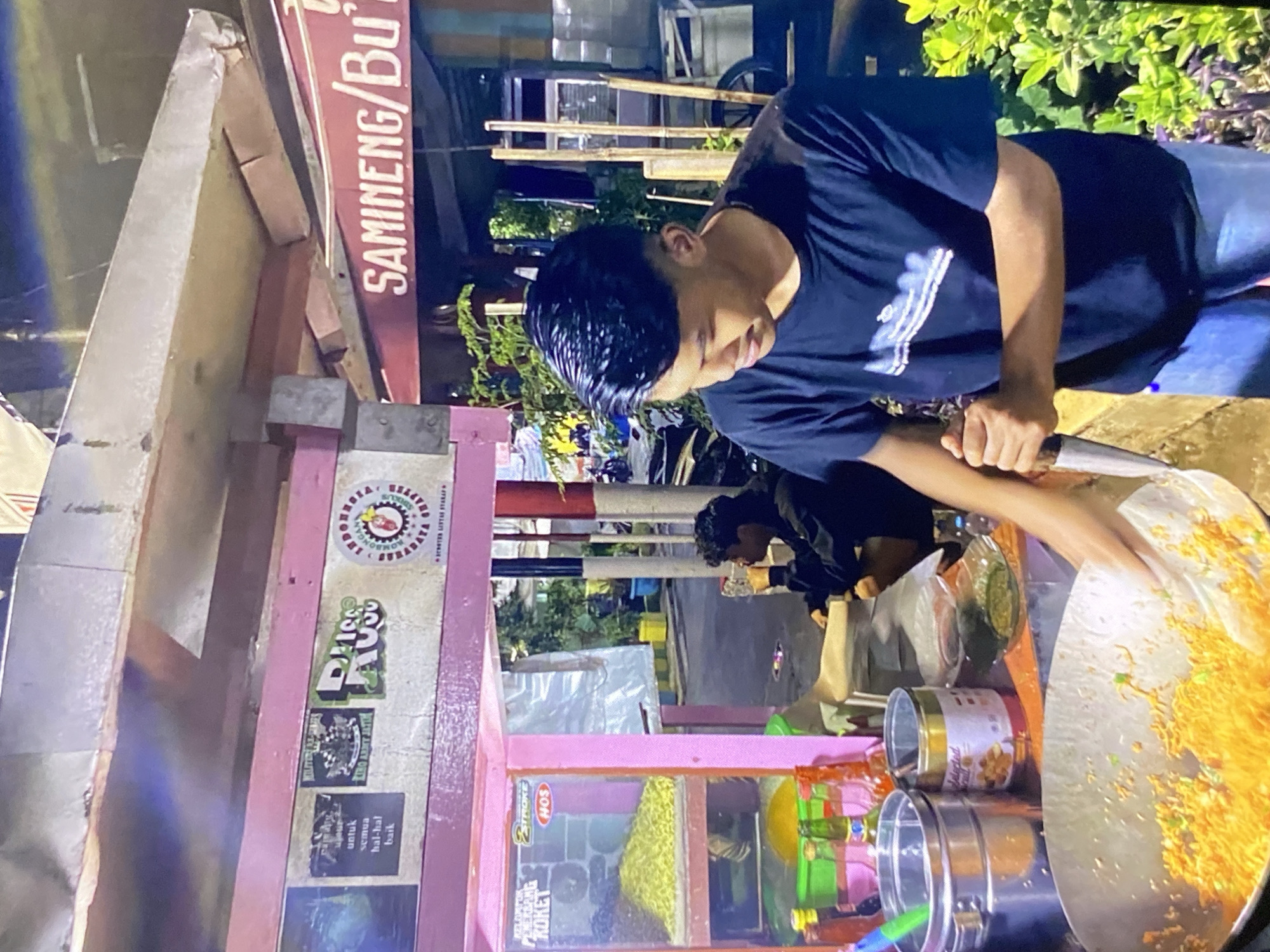 In the city of Tebel, Indonesia, a young man is captured in a low-angle medium shot as he prepares food at an outdoor stall. The stall features a bright pink facade with various decals and posters, including one that reads "RUSI". To the right, a red awning displays the text "SAMINENG / BU'T". The foreground is dominated by the man wearing a dark blue t-shirt and light blue jeans, actively stirring a wok filled with orange noodles and other ingredients. His right hand grips a utensil, and his focus is directed downwards at the cooking process. Behind him, another person sits at a table, seemingly observing or waiting. The background suggests a street-side setting at night, with blurred lights and vegetation. The stall is equipped with containers of ingredients and bottles, indicating it is an active food vendor.