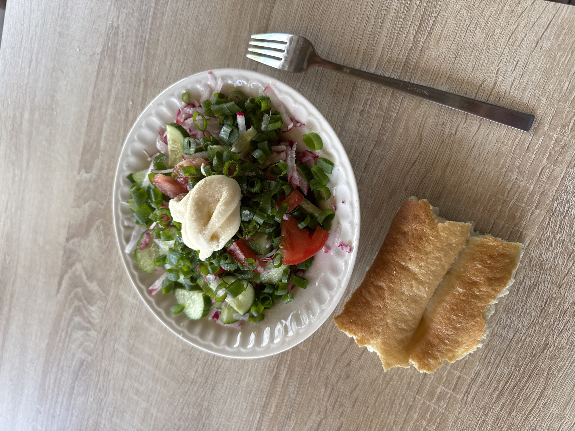A top-down shot shows a white plate containing a salad composed of sliced green onions, cucumbers, radishes, and tomatoes, topped with a dollop of a light-colored sauce. A silver fork lies next to the plate, and a piece of torn bread is adjacent to the salad. The setting is likely a tabletop with a light, wood-grain finish. The location is Chaikovsky, Russia. No people are visible in the image.
