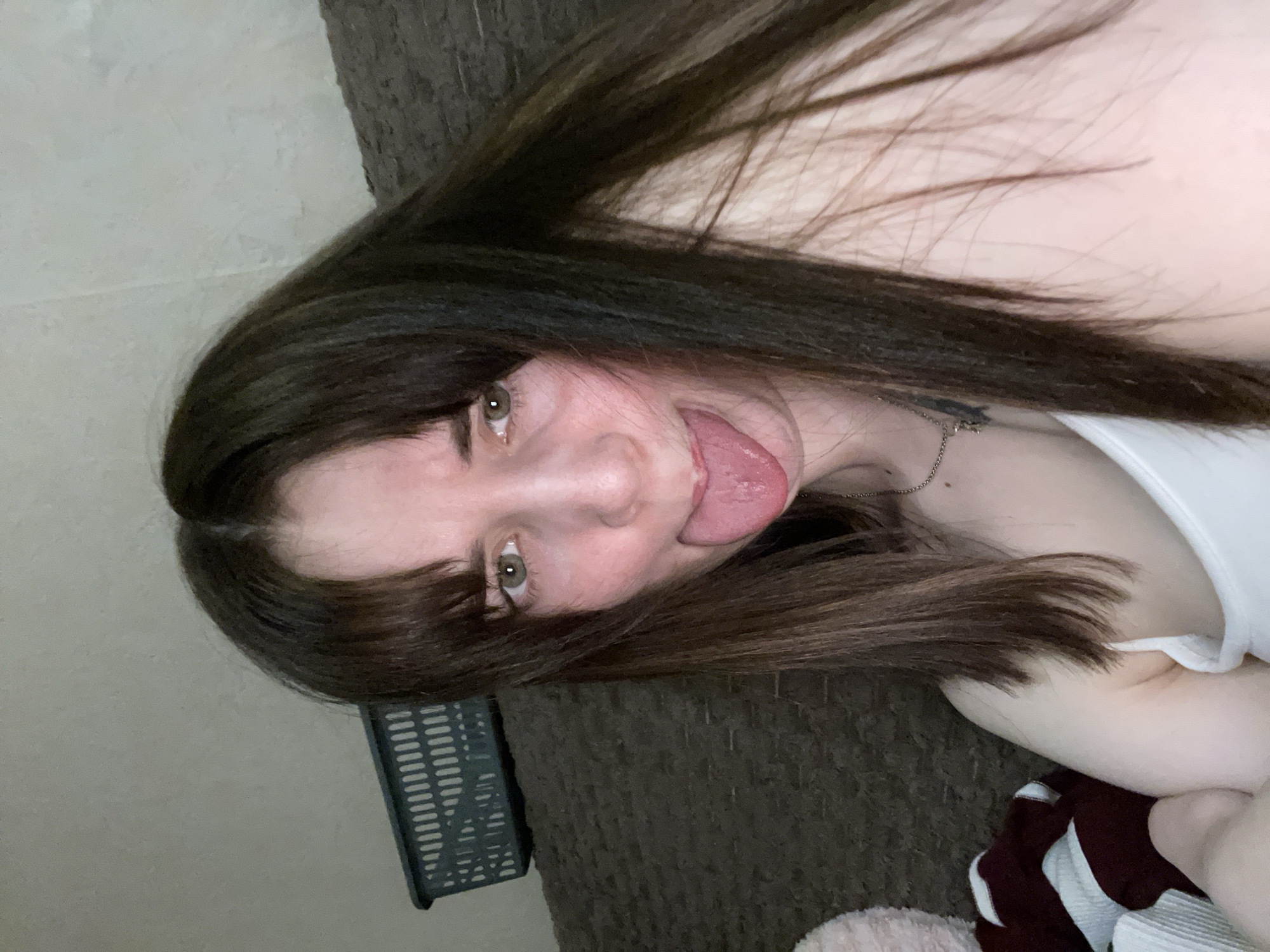 A young woman with long brown hair is sticking her tongue out at the camera. She has fair skin and green eyes. She is wearing a white tank top and a silver necklace. She is lying on a brown blanket. A black plastic basket is visible in the lower left corner of the image. The location is unknown and there is no verifiable evidence that would suggest this photo was taken in Chaikovsky, Russia.