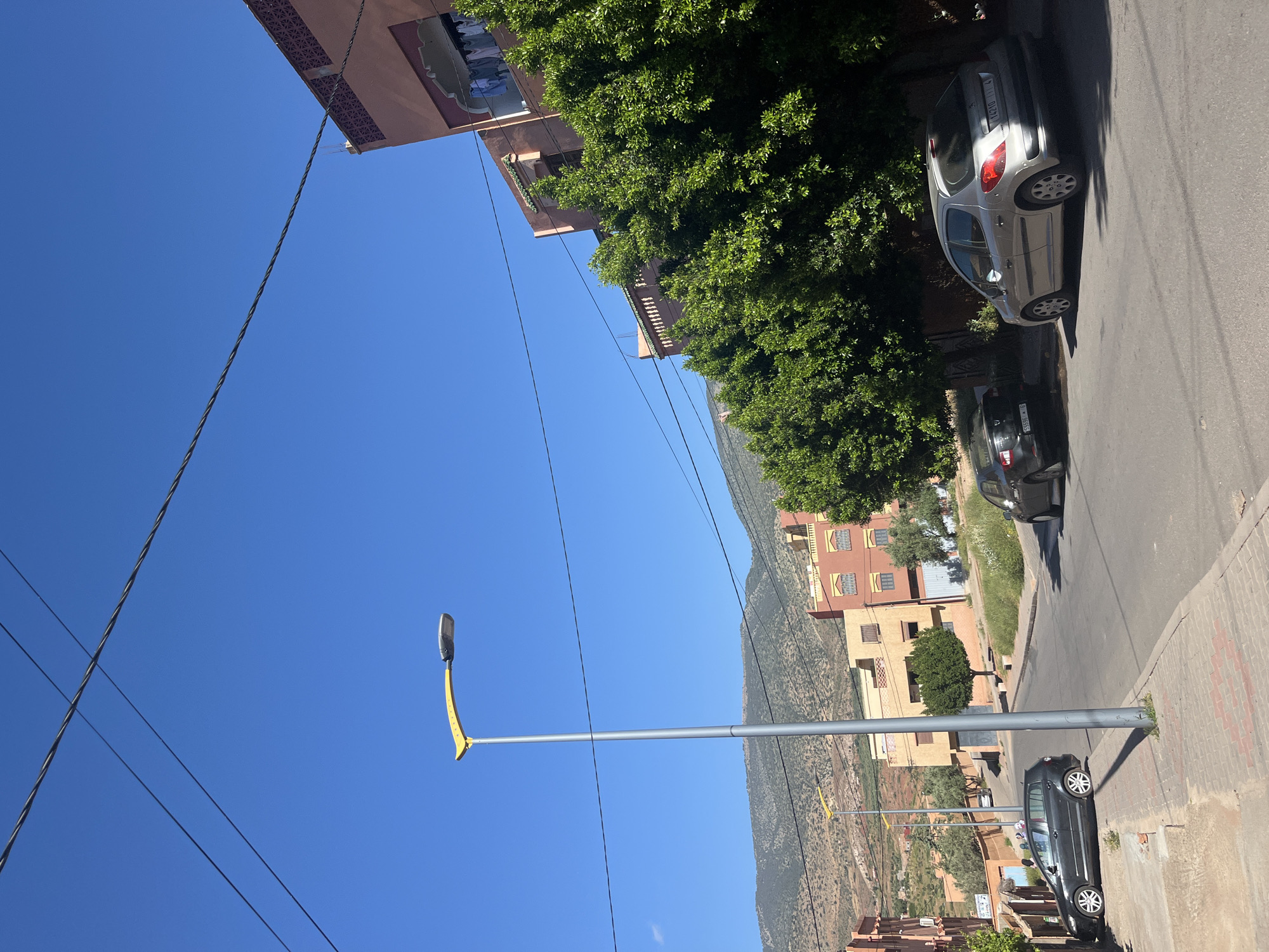 The image depicts a street scene in Ouaouizert, Morocco, under a clear blue sky. A street lamp with a yellow curved arm extends into the frame. Overhead electrical wires crisscross the sky. Buildings, ranging in height and color from peach to yellow, line the street and reach up into the tree-covered hillside in the background. Several cars are parked along the side of the street. The road is paved and bordered by a concrete curb.