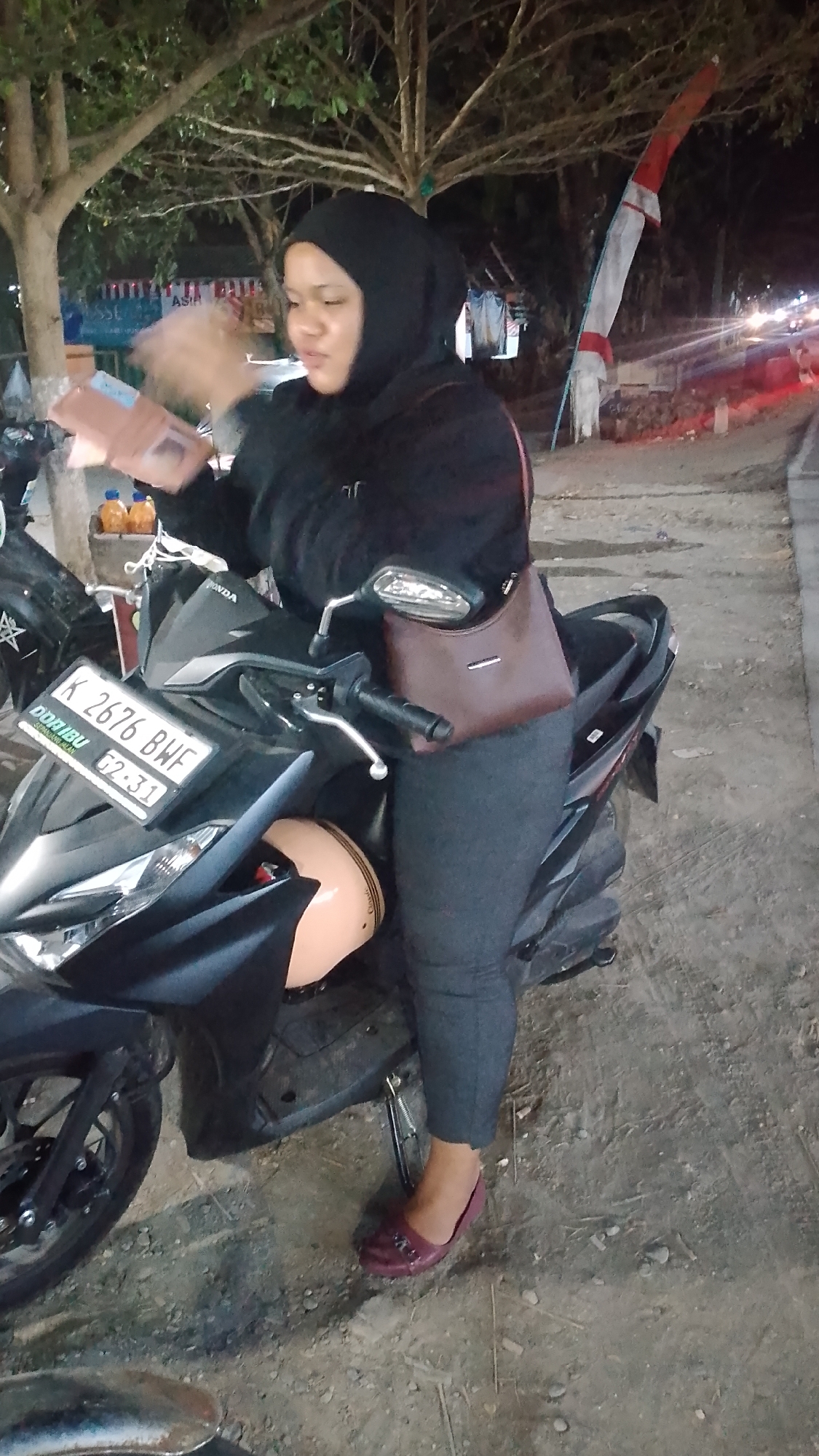 A woman wearing a black hijab and dark grey leggings stands next to a black scooter in what appears to be Karanglangu, Indonesia. She is holding a pink wallet or small book in her hands, and her left hand is blurred, suggesting movement. The scooter has a license plate that reads "K 2676 BWF". A light brown helmet is visible on the scooter. The background shows trees with sparse leaves, a red and white flag on a pole, and distant streetlights, indicating it is nighttime. The ground is a mix of dirt and gravel.