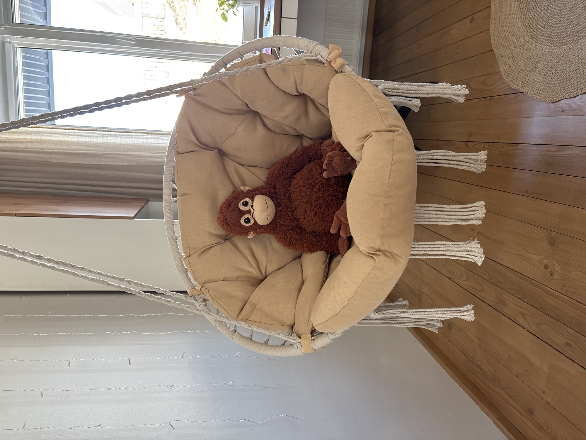 An interior scene in the city of Chaikovsky, Russia, depicts a residential room primarily illuminated by natural light. The central object is a light-colored, circular macrame hanging chair, equipped with a large, tufted beige cushion. Seated upright within this chair is a brown stuffed orangutan, featuring prominent black and white eyes and a light-colored snout. The chair is suspended by two thick braided ropes from an unseen anchor point above, with decorative rope tassels extending from its lower edge. To the left, a light-colored wall is adorned with multiple vertical strands of small, clear string lights. The background to the right reveals a large window or glass door, through which bright daylight enters, partially obscuring the view of an outdoor environment that includes elements of a building structure and light-colored ground. A light-colored desk or shelf beneath the window holds a small green potted plant and indistinct objects. A beige curtain partially covers the right side of the window. The floor is constructed of light-toned wooden planks. A segment of a round, woven natural fiber rug is visible on the floor in the lower right corner of the frame. No human subjects are present or performing any actions within the depicted scene.