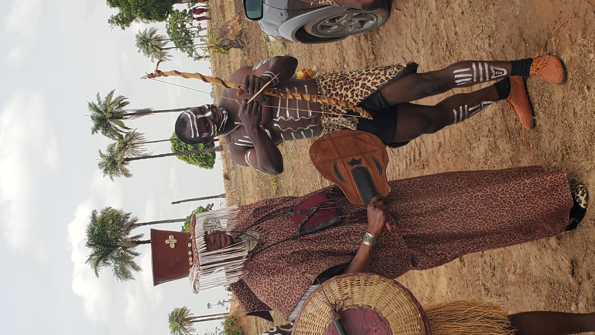 Two individuals are prominently featured in this outdoor scene, with palm trees visible in the background, suggesting a tropical or subtropical environment. The location is Akwanga, Nigeria. The individual in the foreground wears a brown leopard-print garment and a headdress adorned with white hanging strands and embellishments. They hold a brown fan-like object. Beside them is another individual, painted with white markings on the face and body, and wearing a leopard-print skirt, brown shoes, and holding a bow and arrow. The ground appears to be sandy or earthy. A vehicle is partially visible in the upper part of the image.