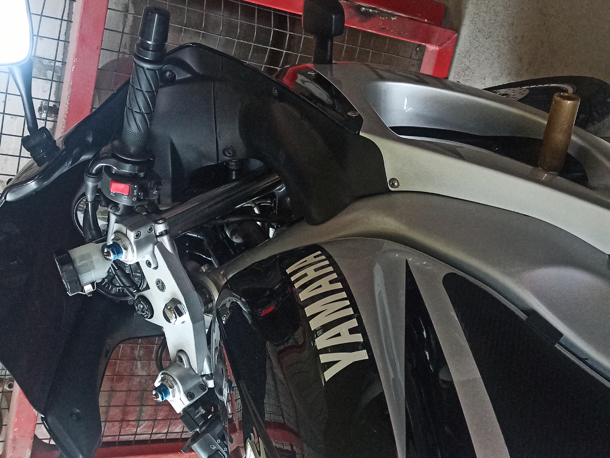 The image depicts a Yamaha motorcycle, primarily showcasing the front section. Visible elements include the handlebars with controls, the front suspension components, parts of the fairing with the "YAMAHA" logo, and a glimpse of the front wheel. The motorcycle is parked on a metal rack. The environment seems to be an indoor space, possibly a garage or a workshop. The city and location are not discernible from the image.