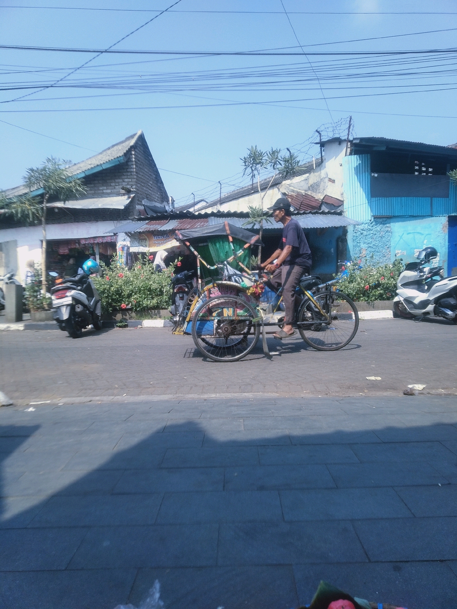 The image depicts a street scene in Cianjur, Indonesia, under a clear blue sky with overhead power lines. In the foreground, a man wearing a dark t-shirt, dark pants, and a cap is pedaling a becak, a three-wheeled passenger bicycle, on an asphalt road. The becak has a green canopy and is adorned with colorful decorations. To the left of the becak, a silver scooter with a blue helmet is parked. Behind the scooter, a building with a corrugated metal roof and a light-colored facade is visible, with potted plants and flowering bushes in front of it. Further in the background, there are other buildings, including one with a vibrant blue facade, and a white scooter is parked. The overall impression is of a bustling urban environment with a mix of traditional and modern transportation.