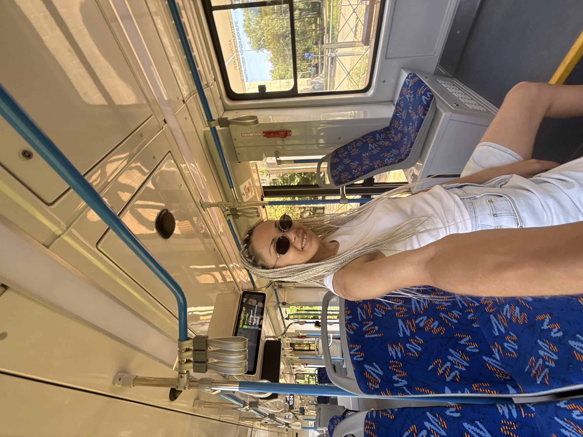 A light-skinned woman with blonde braided hair and sunglasses is taking a selfie inside a public transport vehicle, possibly a bus or tram, in Rusanovo, Russia. She is wearing a white tank top and denim shorts. The interior of the vehicle is visible, featuring blue seats with an orange and white pattern, a blue handrail, and a digital display showing route information. Outside the window, trees and buildings can be seen. A sign on the window reads, "ПОЖАЛУЙСТА, НЕ ОТКРЫВАЙТЕ ФОРТОЧКИ ПРИ РАБОТАЮЩЕМ КОНДИЦИОНЕРЕ" which translates to "PLEASE DO NOT OPEN THE WINDOWS WHEN THE AIR CONDITIONER IS ON."