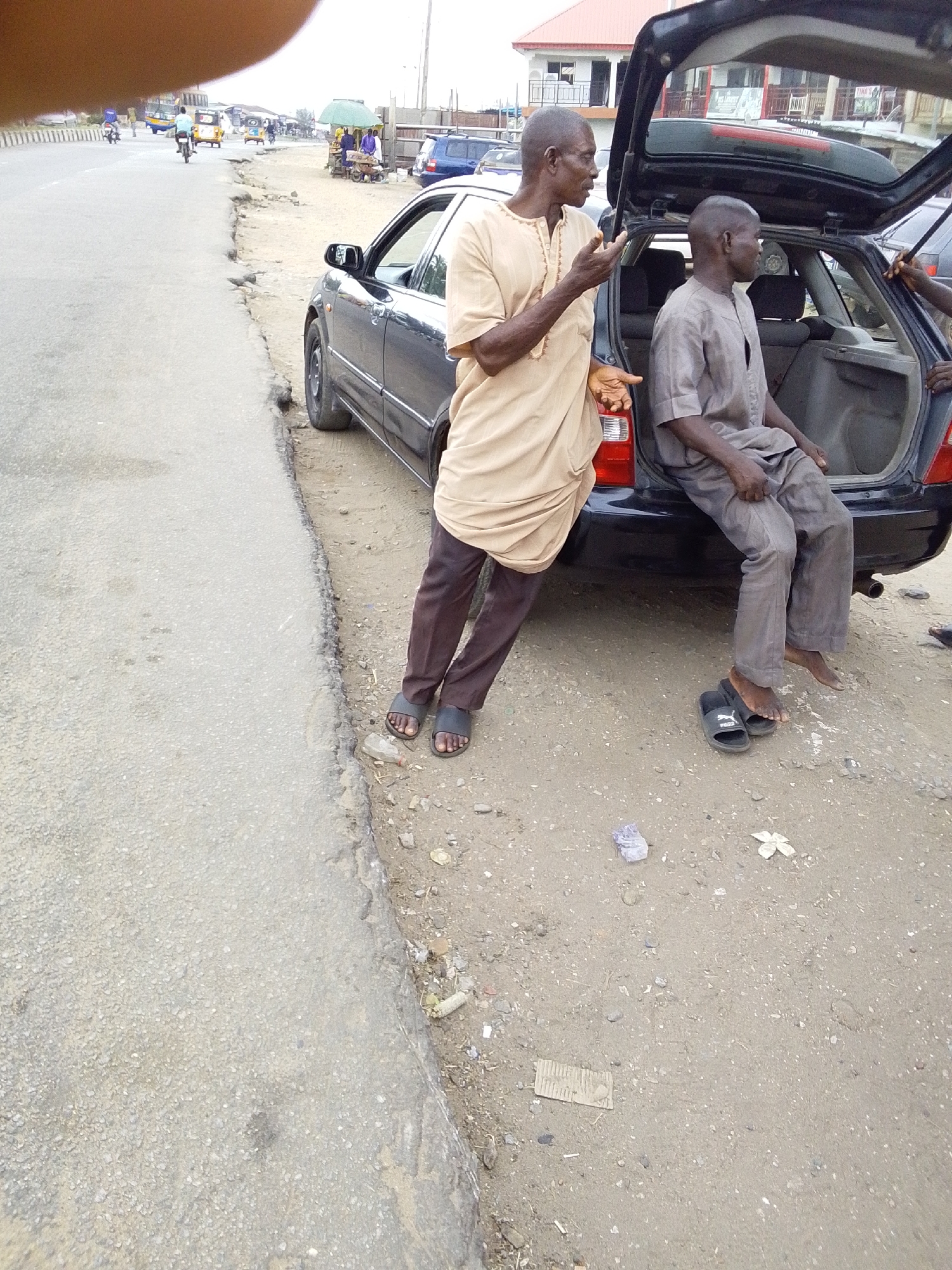 Two middle-aged African men are beside a black car with its trunk open on a dirt shoulder adjacent to a paved road. One man is standing, wearing a long tan shirt and dark pants, gesturing with his right hand. The other man is sitting on the edge of the open trunk, wearing a gray shirt and pants. Both men are wearing sandals. The location appears to be Lokoja, Nigeria, evidenced by the style of dress and the architecture in the background. A building with a red roof can be seen in the background. The ground around the car is dirt and scattered debris. There are other vehicles and pedestrians on the road in the distance.