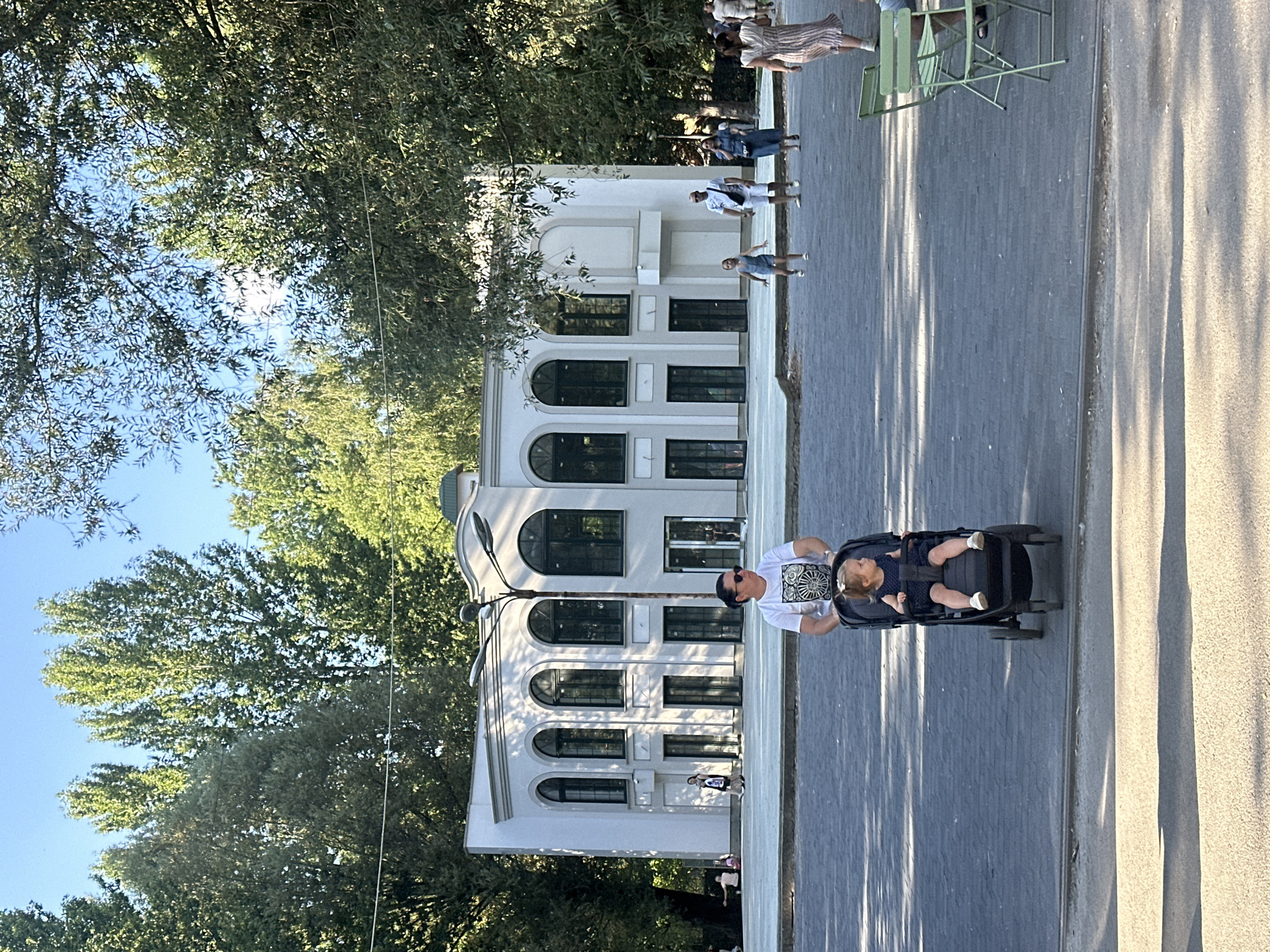 The image depicts a sunny day scene in Ternopil, Ukraine, specifically at the "Podoliany" park area. A woman is seen pushing a stroller with a young child in it. In the background, a modern white building with arched windows stands prominently. Several other individuals are visible walking around the park. The ground is paved with dark gray cobblestones, and lush green trees surround the area, creating a pleasant park atmosphere. A streetlamp is positioned in the middle ground. The lighting suggests it is daytime, with the sun casting shadows on the ground.