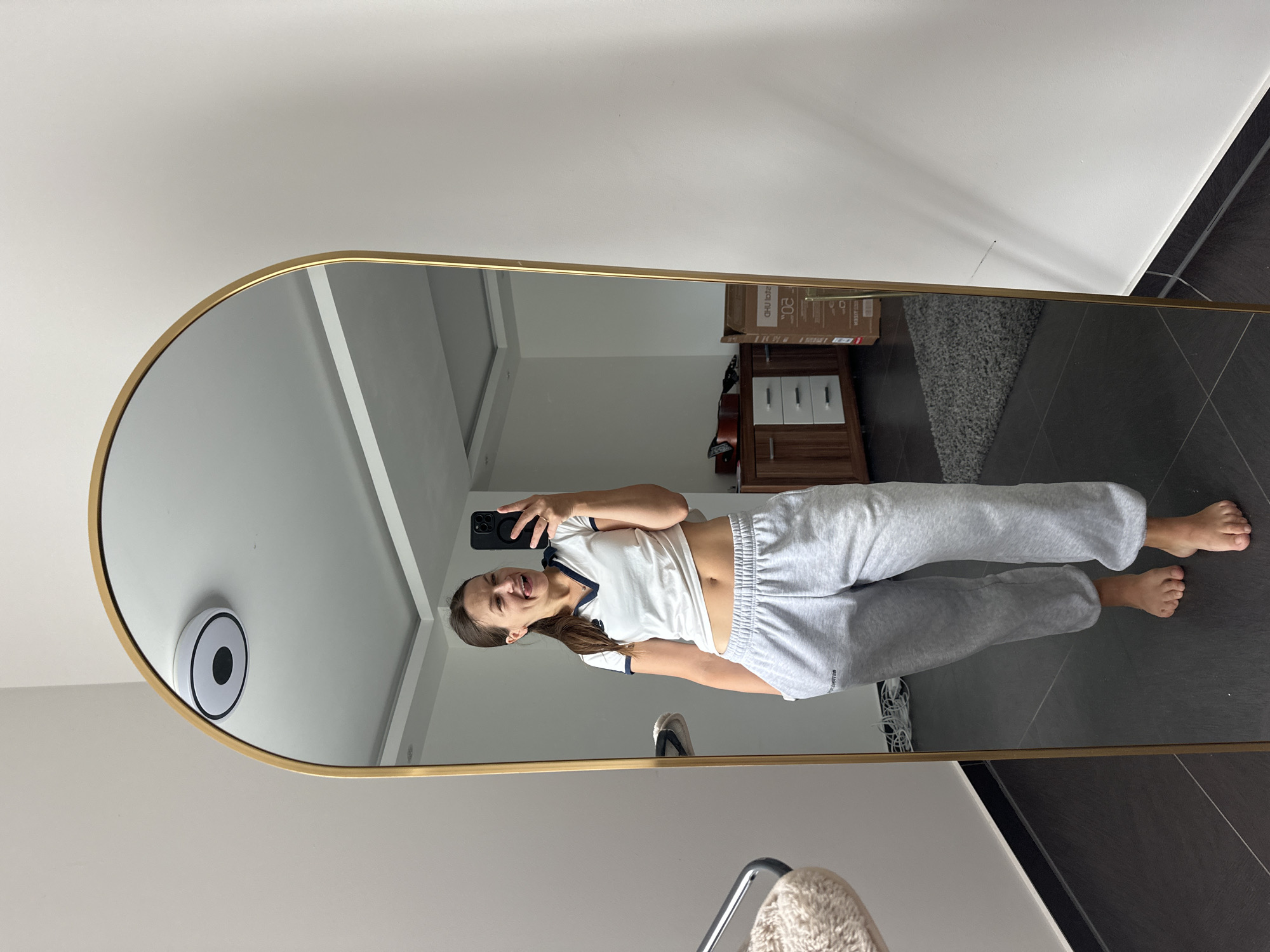 A woman is taking a selfie in a full-length mirror in Kutenholz, Germany. She is wearing a white t-shirt with a navy trim and grey sweatpants. Her left hand is in her pocket and her right hand is holding her phone. Her tongue is sticking out and her eyes are closed. The mirror is framed in gold and has an arched top. In the background, there is a dresser with drawers, a grey rug, and a cardboard box. The floor is tiled and dark. The walls are white. A white circular object with a black center is affixed to the ceiling.