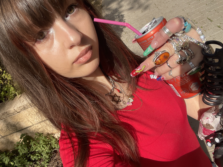 A close-up, high-angle self-portrait features a young individual with long brown hair, reddish highlights, and bangs, looking directly at the camera. She wears a red top and numerous pieces of jewelry, including multiple necklaces with pendants such as a Star of David and a red Hamsa/Evil Eye charm, along with several rings on each finger, including a large amber-colored stone ring on her right ring finger. Her long fingernails are elaborately decorated with various designs, including a green serpentine pattern and dice motifs. She holds two red Coca-Cola cans, visible with Hebrew text; one contains a pink plastic straw. Partially visible near her left side is a floral-patterned fabric, and a black hair claw clip is near her right hand. The background indicates an outdoor setting with green foliage and a light-colored paved surface under bright sunlight, consistent with an urban environment in Raanana, Israel, as evidenced by the Hebrew branding on the cans.