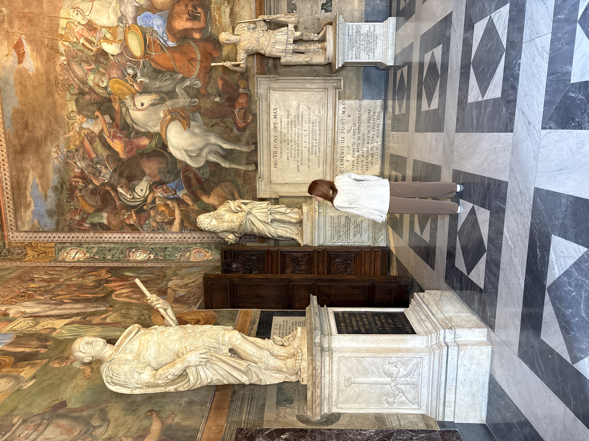 The image shows the Sala dei Pontefici (Hall of the Pontiffs) in the Capitoline Museums, Rome, Italy. A woman in a white jacket and brown pants is looking at the marble tombs and statues displayed along the walls. A large fresco depicting a battle scene covers the upper part of the walls. Marble flooring with a geometric pattern is visible. Latin inscriptions are inscribed on the tombs.