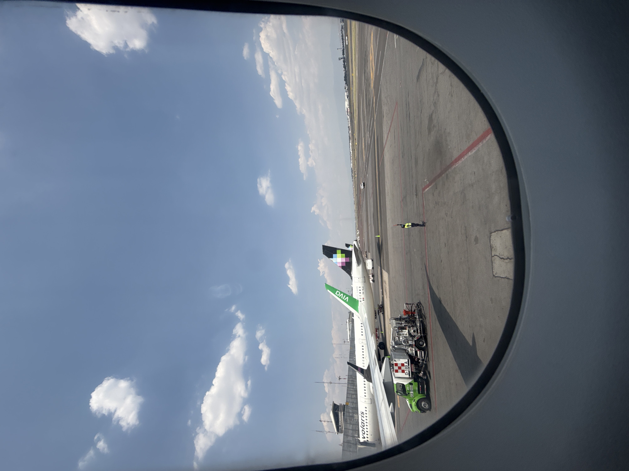 Captured from an airplane window, the image depicts a sunny day at an airport tarmac, with a clear blue sky and scattered white cumulus clouds. The curved outline of the aircraft window frames the scene.
Two passenger jets are visible on the tarmac. The prominent aircraft in the foreground is a white jet with "volaris" written on its fuselage and "VIVA" in green on its tail, which also features a distinctive black and green geometric pattern. Partially visible behind it is another white aircraft, whose tail is black with multi-colored geometric shapes and includes text like "CIA" and "DXA".
A green and white ground support vehicle, marked with "D-189 A" and "ASA 4104", is positioned next to the wing of the Volaris aircraft. The concrete tarmac below is marked with various yellow and red lines. In the distance, a person wearing a yellow high-visibility vest stands on the tarmac, appearing to direct ground operations with outstretched arms. Further in the background, airport buildings and a faint mountainous or urban landscape are visible.
The scene captures typical daytime airport activity, suggesting preparations for departure or arrival. The presence of Volaris and Viva Aerobus aircraft strongly indicates an airport in Mexico, aligning with the provided location context of Mexico City, Mexico.