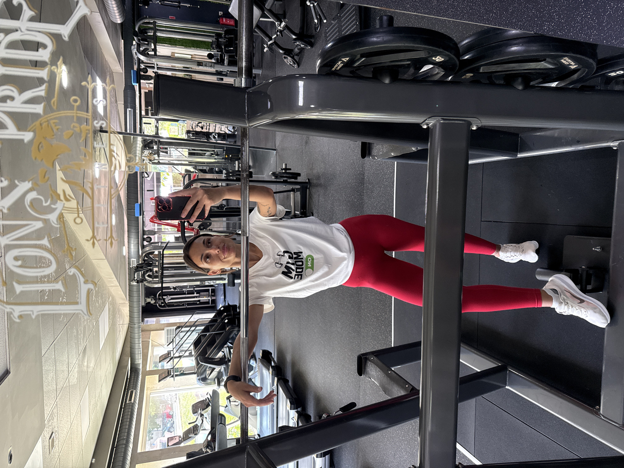 In the city of Pleven, Bulgaria, a woman is captured in a mirror selfie at a gym, reflecting the "Lion Pride Fitness" logo on the ceiling. She stands in front of a weight rack, holding her phone in her left hand and resting her right arm on the barbell. The woman is wearing a white t-shirt with a graphic that reads "GYM MODE" and red leggings, paired with white athletic shoes. The gym environment is visible in the background, featuring treadmills, weight machines, and black rubber flooring. The lighting suggests it's daytime.
