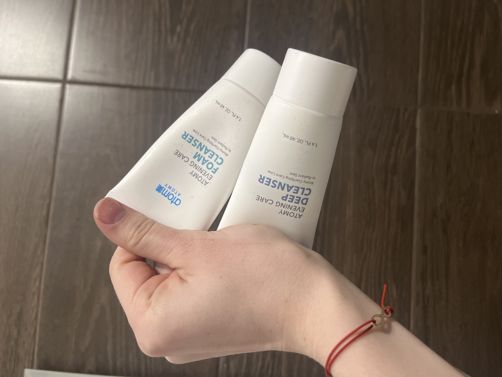 The image shows a hand holding two tubes of Atomy cleanser products. The tube on the left is "Atomy Evening Care Foam Cleanser" and the tube on the right is "Atomy Evening Care Deep Cleanser". Both tubes are white and indicate a size of 1.4 fl. oz. 40 mL. The background is a wall with dark brown rectangular tiles. The hand holding the products has fair skin. A red string bracelet is on the wrist.
