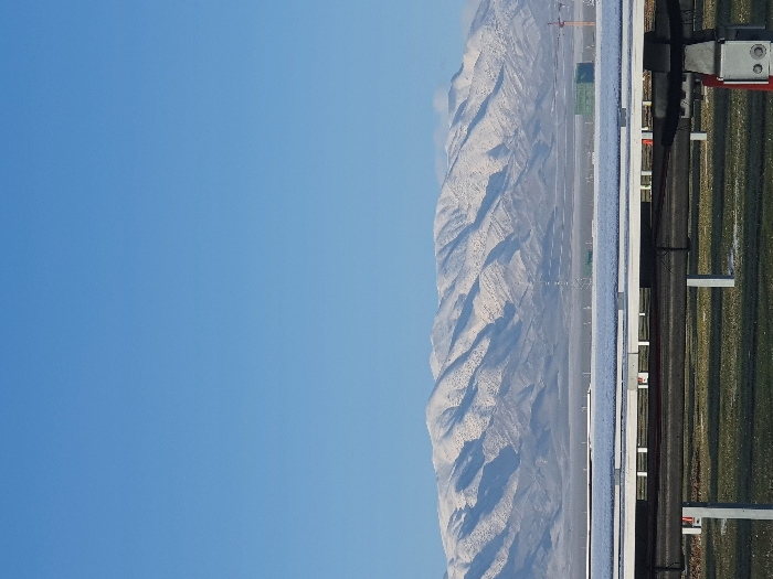 A landscape view features a clear, light blue sky occupying the left portion of the frame. Moving right, a mountain range, dusted with snow, stretches towards the background. In the foreground, a fence or barrier of some sort runs vertically, with linear elements and supports visible against a backdrop of green vegetation. The scene presents a blend of natural and man-made structures.