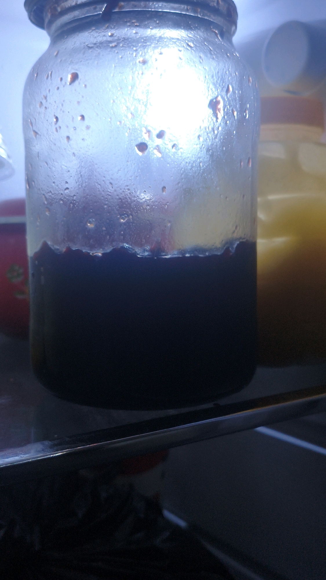 A close-up, low-angle shot captures a glass jar filled with a dark purple liquid, likely jam or preserves, sitting on a shelf. The jar is covered in condensation, with small water droplets clinging to the glass. The lid is a rusty metal screw-top. In the background, another jar with a yellow substance is visible. The scene is dimly lit, with a bright light source from above the jar creating a glare. The foreground shows the edge of a metallic shelf. The image does not provide any specific details about the city of Mehnatabad, Uzbekistan, or any people present.