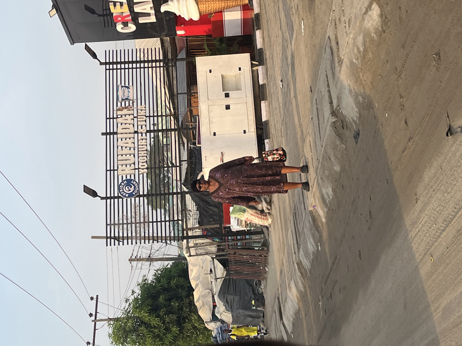 An adult female, dark complexion, stands centrally on a dirt and concrete roadside in Lekki, Nigeria, looking towards the viewer. She wears a long-sleeved, V-neck brown dress with a subtle pattern, dark sunglasses, and dark flat shoes. She holds a floral-patterned tote bag in her right hand. To her left, another individual in a bright yellow top walks away from the camera. The road exhibits tire tracks and is bordered by a concrete kerb and gutter. A small bird is visible on the road surface near the bottom.
Behind the woman, a large white industrial generator, labeled "MIKANO," rests on a raised platform. To the generator's right, a dark commercial structure features partially visible "CREAM LAND" lettering and a large, decorative ice cream cone sculpture. Further left, a series of makeshift market stalls or structures, constructed from dark tarpaulins and metal frames, are visible, alongside piles of bundled materials.
In the background, a large billboard with horizontal black bars displays "RUFAIDAH YOGHURT & FURA" in white text, accompanied by a yogurt cup logo. Overhead, power lines crisscross a clear, pale blue sky. Green foliage from trees is visible behind the market stalls and below the billboard.