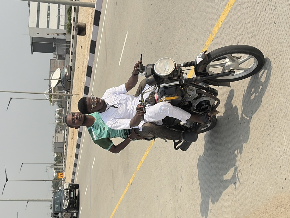 Two adult males are riding a black motorcycle on a multi-lane paved road in Alaguntan, Nigeria, under bright daylight conditions. The driver, a light-skinned male wearing a white short-sleeved traditional outfit, dark-framed glasses, and dark flip-flops, grips the handlebars. The passenger, a darker-skinned male in a green and black athletic shirt and dark trousers, sits behind the driver, looking forward. The motorcycle has a round headlight and silver five-spoke wheels. The road surface is light-colored, with a solid yellow line on the left and a black-and-white striped curb marking a median to the right. Tall streetlights with flat heads line the road. In the background, a dark SUV travels in the opposite direction on the far side of the road. Further in the distance are modern buildings, a large satellite dish structure, a visible "PETROTECH" sign, and another car. Strong shadows are cast on the road surface, indicating direct sunlight.