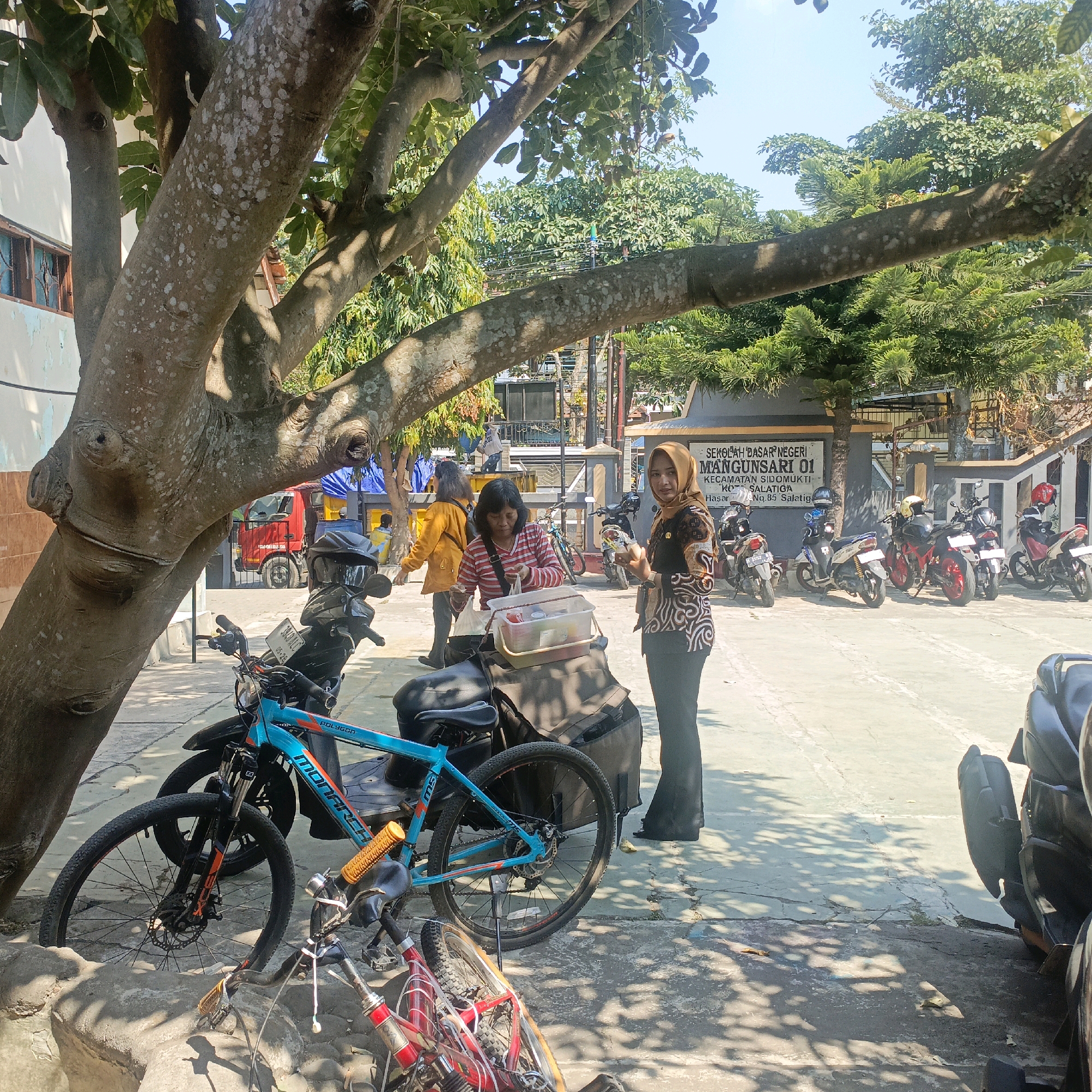 In Salatiga, Indonesia, a group of people are gathered outdoors near a school. In the foreground, a blue bicycle with the brand name "Polygon" is parked, and behind it, a portion of a scooter is visible. Further back, a woman in a striped shirt and a headscarf is interacting with a container on the back of a parked scooter. To her right, another woman in a patterned top and headscarf is holding a small white object. Behind them, several motorcycles are parked in front of a building with a sign that reads "SEKOLAH DASAR NEGERI MANGUNSARI 01." A red truck is also visible in the background. The scene is set under the shade of a large tree, with dappled sunlight falling on the pavement and objects.