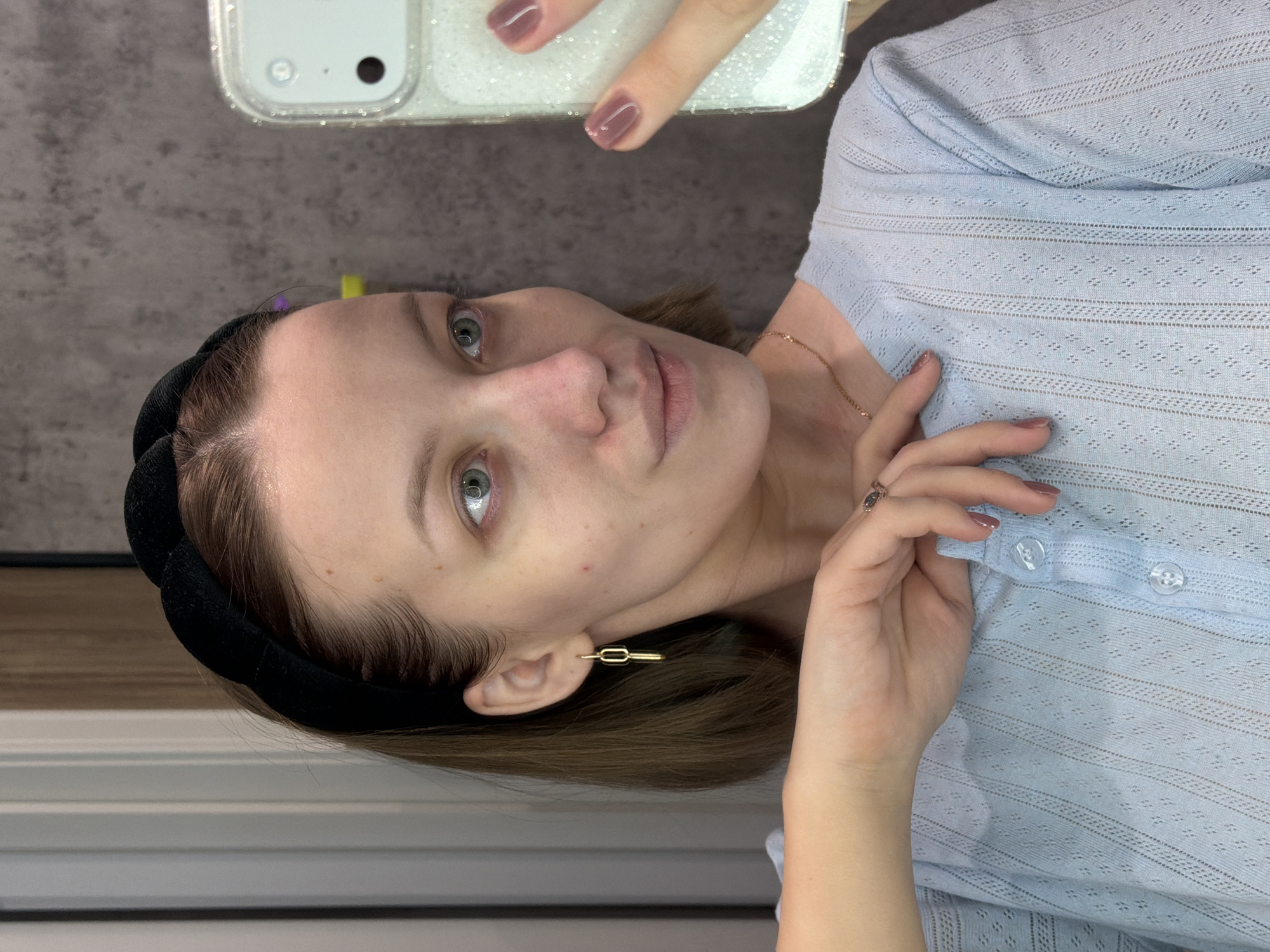 A young woman in Reshetnikovo, Russia, is captured in a selfie, with the image rotated 90 degrees counter-clockwise. She is holding a smartphone with a clear, glittery case in her right hand, visible at the top, as she takes the photograph. Her green eyes are focused directly on the camera, and she has a neutral, composed expression.

She wears a light blue, short-sleeved top featuring an intricate eyelet knit pattern with horizontal bands and small diamond-shaped perforations. Her light brown hair is styled back with a wide black velvet headband. Adorning her left ear is a modern, gold-toned geometric bar earring, and a delicate gold chain necklace is visible around her neck. On the ring finger of her left hand, which rests near her chest, she wears a ring with a dark stone. Her fingernails are painted a muted purplish-brown.

The background suggests an indoor setting, appearing as a dark gray, textured wall, possibly concrete or painted plaster. The lighting is soft and even, characteristic of indoor illumination. There is no visible text in the image.