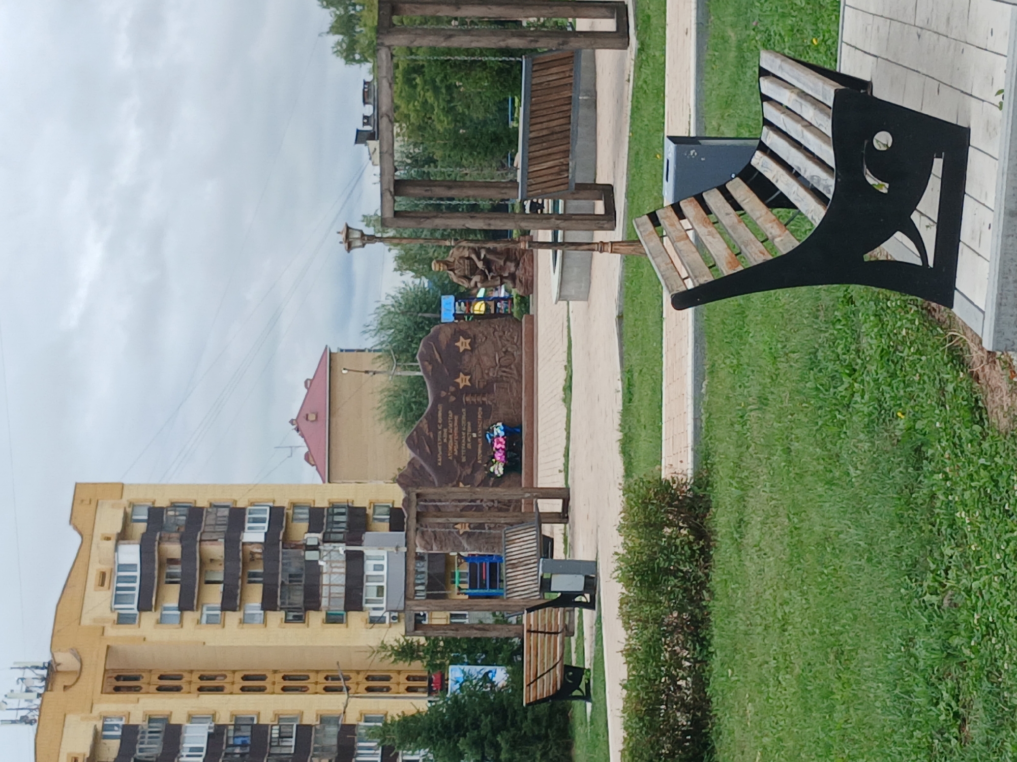 The image depicts a public park area in the city of Shchuchinsk, Kazakhstan, featuring several key elements. Dominating the background is a tall, multi-story apartment building with a yellow facade and numerous balconies. Adjacent to it, a smaller, reddish-brown building with a pitched roof is visible. In the mid-ground, a memorial monument stands prominently, consisting of a large, textured stone slab adorned with reliefs and stars, accompanied by a statue of a seated figure. In front of the monument, a wooden swing set with a bench-like seat is present. Several park benches are strategically placed throughout the scene, including a distinctive black and wooden bench in the foreground. Lush green grass and manicured bushes form the landscaping, with paved pathways indicating pedestrian areas. The sky is overcast with gray clouds, suggesting a temperate climate. No individuals are actively present in the immediate vicinity of the memorial or benches, though the presence of the apartment building implies a residential area. The overall scene conveys a sense of quiet public space, likely a memorial park or a communal area within a residential complex.
