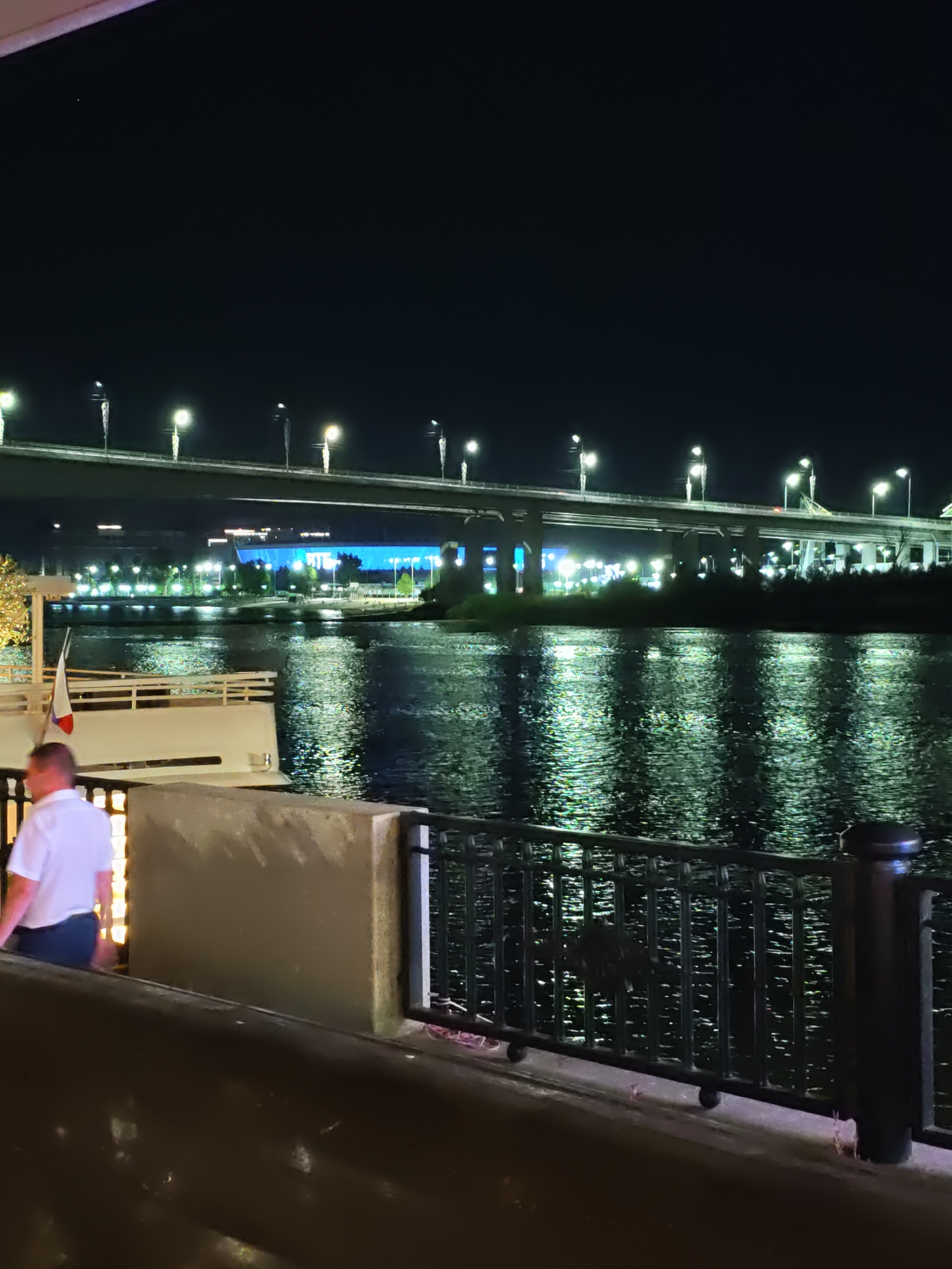An outdoor nighttime scene shows a man in a white polo shirt and dark pants standing on a concrete walkway next to a body of water. In the background, a large bridge with numerous lights spans across the water, with illuminated buildings and trees visible beyond. A white boat with a Russian flag is docked on the left side of the walkway. The water reflects the lights from the bridge and buildings, creating a shimmering effect. The scene is likely the embankment in Rostov-on-Don, Russia.