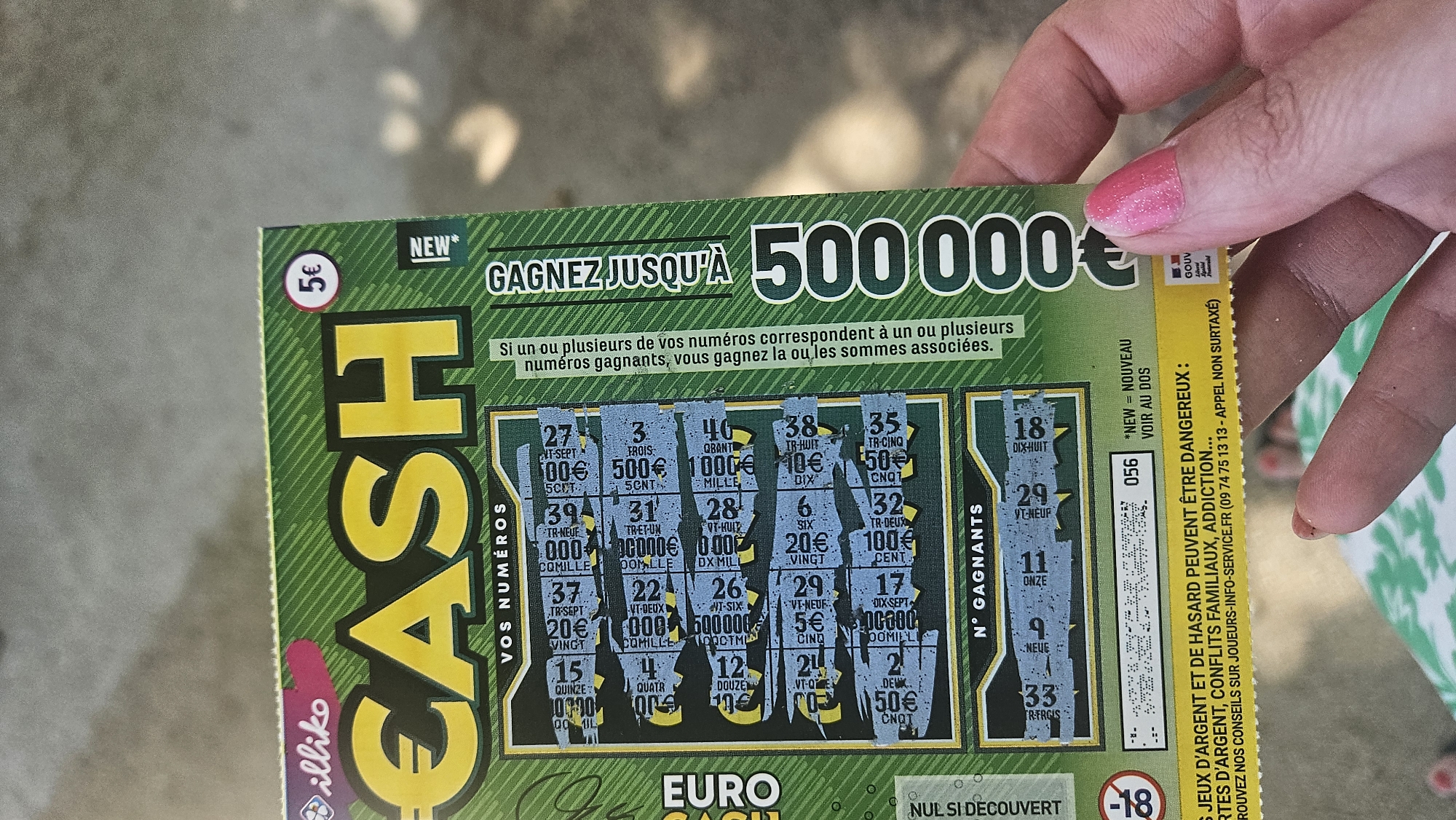 An overhead, close-up shot features a hand with bright pink fingernail polish holding a scratch-off lottery ticket. The ticket is predominantly green and yellow and features the word "CASH" in large, bold, yellow letters across the top. Below "CASH" are the words "VOS NUMÉROS" (Your Numbers) and a section of scratched-off areas. The ticket also displays a large "500 000 €" graphic on the right side. A smaller "5€" is visible in a red circle on the upper right of the ticket, and the word "NEW" is printed in small white letters. The bottom of the ticket contains warnings in yellow text regarding the dangers of gambling and a phone number for information. The background appears to be an outdoor setting, possibly a street or public area, with a textured gray surface and some blurry green foliage. The context of L'Île-Rousse, France, is not directly ascertainable from the image itself, but the "€" symbol indicates it is within the Eurozone.