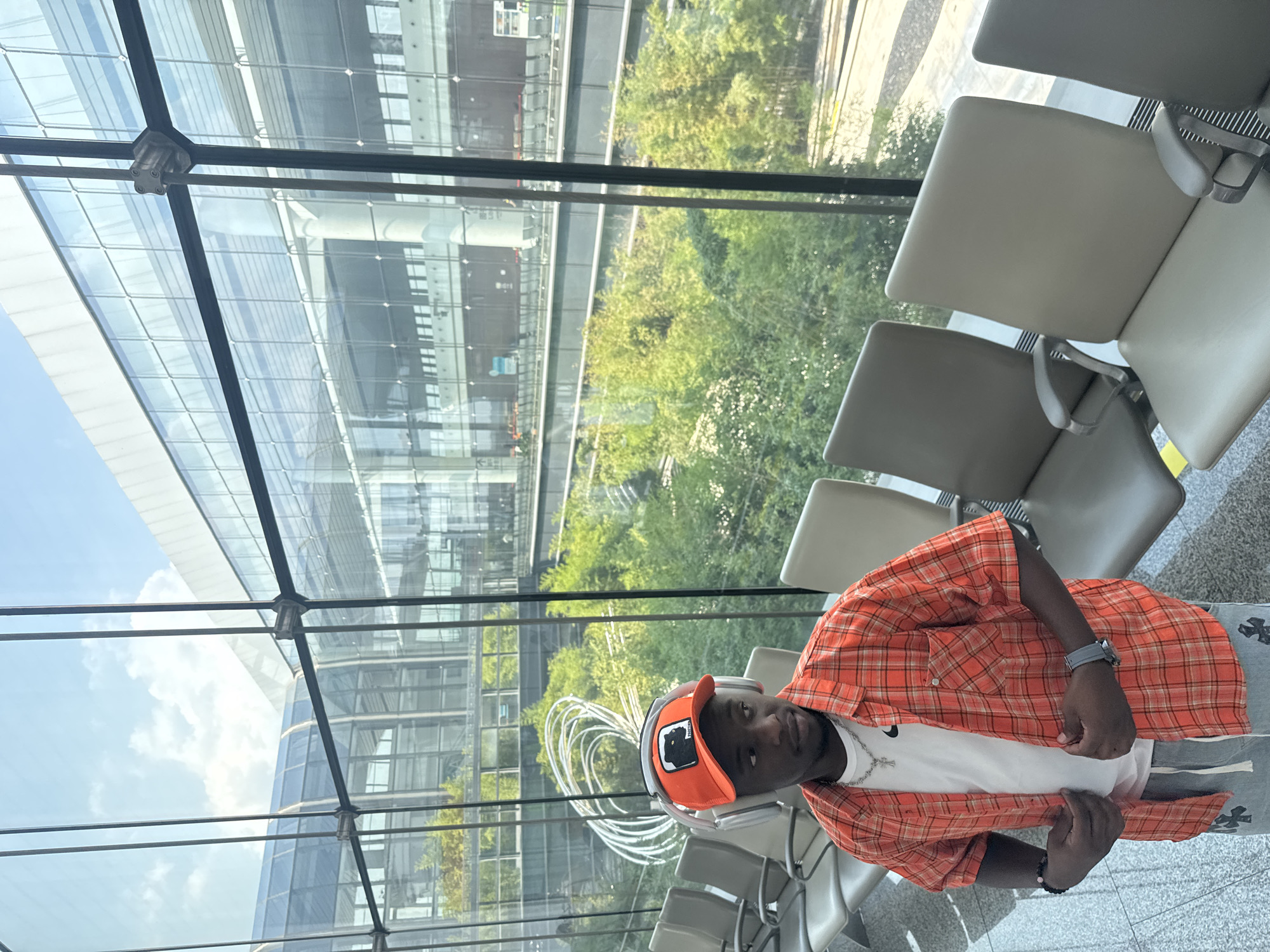 The image captures a young Black male wearing an orange plaid shirt, a white t-shirt, grey sweatpants, and an orange baseball cap with headphones. He is standing in what appears to be an airport waiting area, with rows of empty grey chairs behind him. Large windows overlook a cityscape with a modern architectural building and lush green trees. The reflection in the windows suggests an outdoor environment bathed in sunlight. The man is looking to his right, with his hands on his shirt. The overall scene conveys a sense of travel and anticipation. The mention of Guangyang District, China, in the prompt appears to be external information not directly discernible from the image itself.
