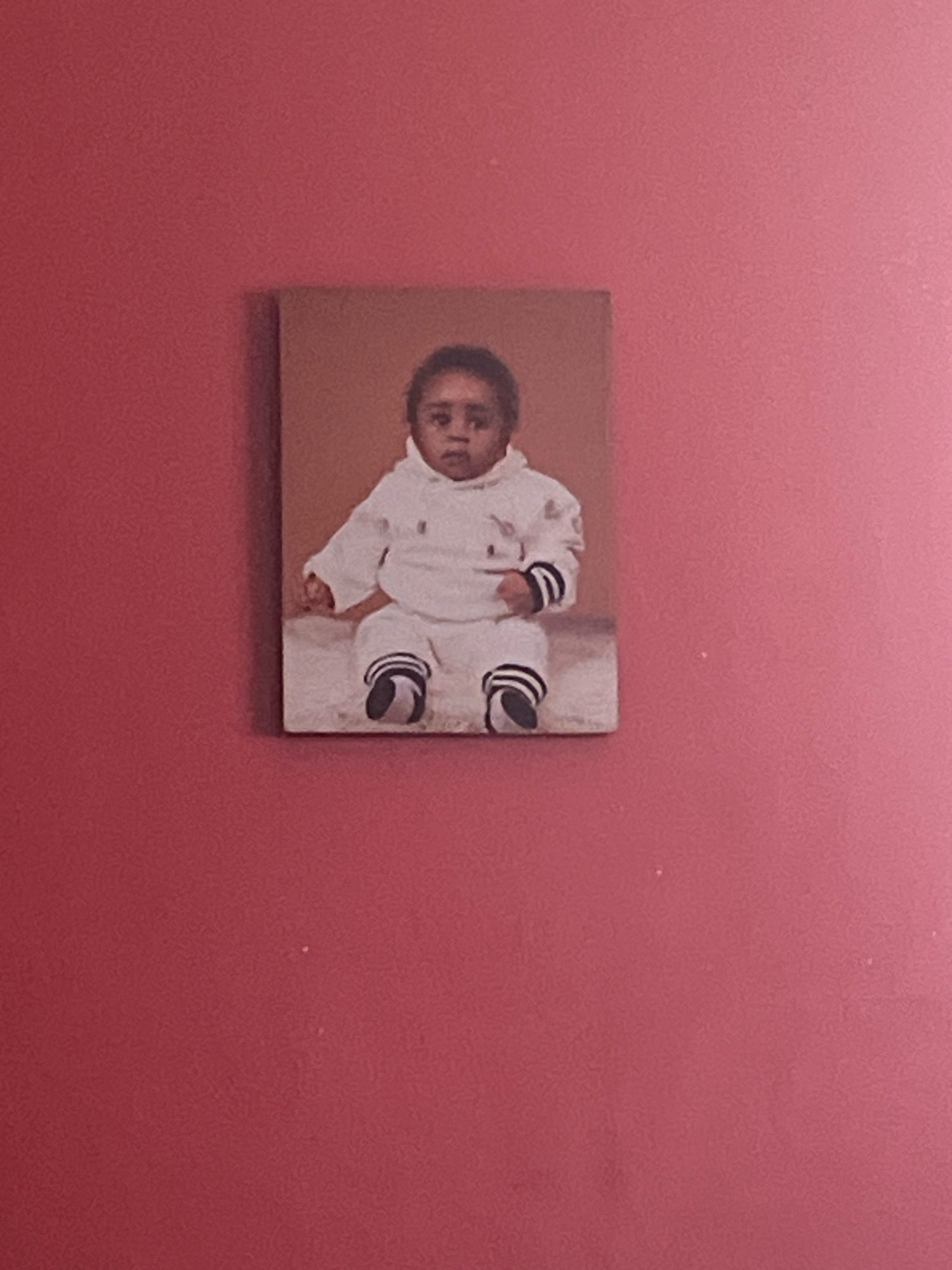 A framed photograph of a baby is mounted on a pink wall. The photograph depicts a young child with dark curly hair, wearing a white hooded outfit with dark trim. The baby is sitting with legs bent and feet turned inward, also adorned with dark-trimmed socks or shoes. The background of the photograph is a muted brown. The wall is painted a vibrant pink and shows some texture. The photograph appears to be centrally located on the wall, with significant empty space above and below it. There are no other objects or individuals visible in the frame. The scene is an indoor setting, likely a home environment. This image does not contain any information that would allow for its location in Jos, Nigeria, or any other specific city.