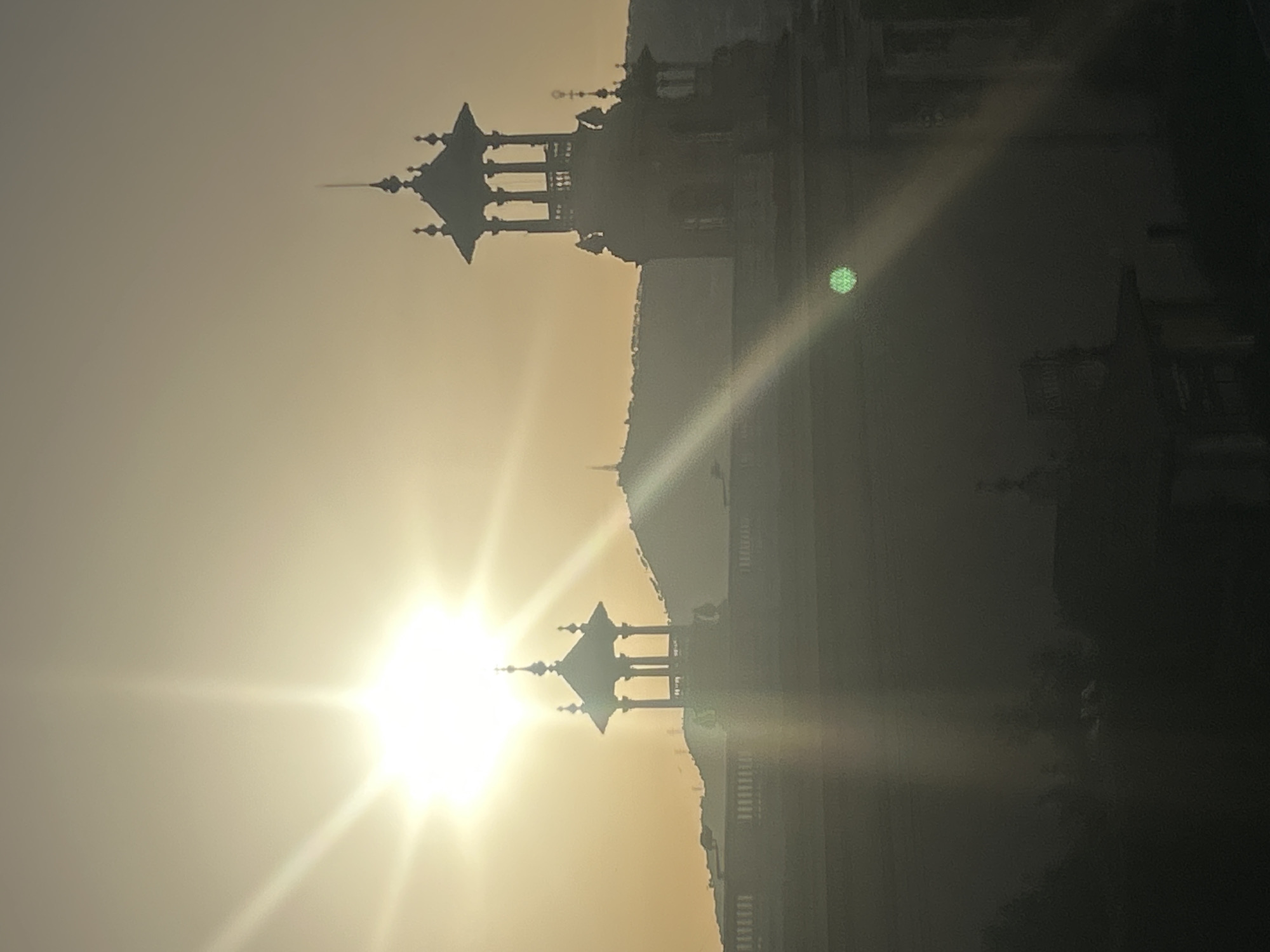 The image depicts a silhouetted view of the Palau Nacional in Barcelona, Spain, against a bright, hazy sky with the sun prominently visible and creating lens flare. The sun is a large, white starburst with radiating beams of light. The Palau Nacional's architecture is visible, including its distinctive towers and rooftop balustrades, all rendered in dark silhouette. A distant, hazy hillside forms the backdrop. The foreground is also in silhouette, obscuring finer details of any structures or vegetation that may be present. The overall impression is one of a bright, possibly late afternoon or early morning sun casting long shadows and creating a somewhat ethereal atmosphere. There are no discernible people or active interactions visible in the frame.