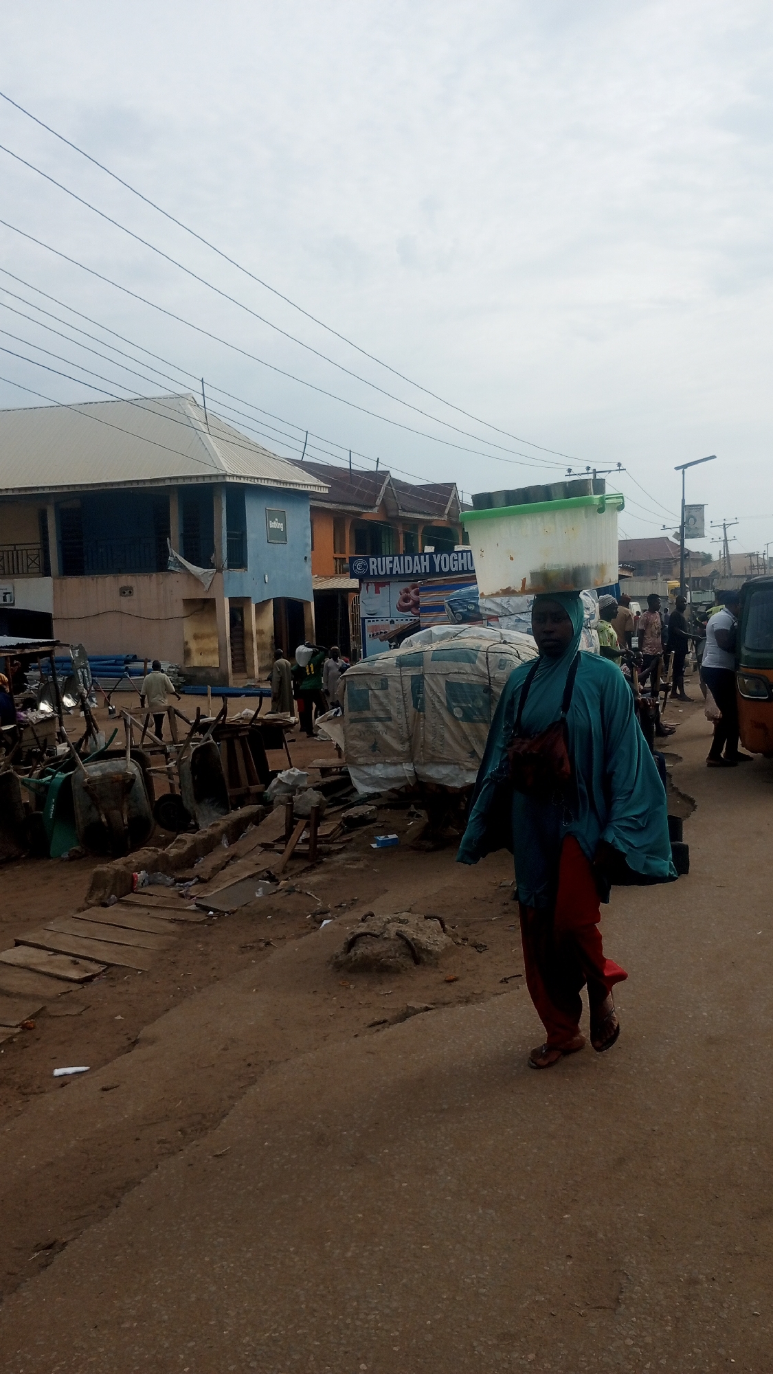 This image depicts a bustling street scene in Ajaokuta, Nigeria, under an overcast sky. In the foreground, a woman walks purposefully towards the viewer, positioned slightly to the right of the frame's center. She wears a teal head covering and a loose-fitting, long teal garment over red trousers, with a small bag slung across her chest. Demonstrating a common local practice, she expertly balances a large, light-colored rectangular container on her head.
The street is a mixture of unpaved, dusty ground on the left, transitioning to a more compact, darker surface in the immediate foreground and where the woman walks. The left side of the street is cluttered with various objects, including several wheelbarrows, stacks of blue pipes, wooden planks, and other materials, some covered with light-colored tarpaulins, suggesting a market or construction supply area.
In the mid-ground and background, numerous other people are visible, walking, standing, and interacting. Many appear to be men dressed in traditional attire. Several multi-story buildings line the street; prominently featured are a light-blue building with a white roof on the left and an orange-brown building further back. Overhead power lines stretch across the cloudy sky.
A clear sign on a shop or stall in the mid-ground displays the text "RUFAIDAH YOGHURT," indicating a business. A small, partially visible yellow auto-rickshaw is seen on the far right, alongside a street light pole. The scene conveys the lively atmosphere of a typical day in a Nigerian town.