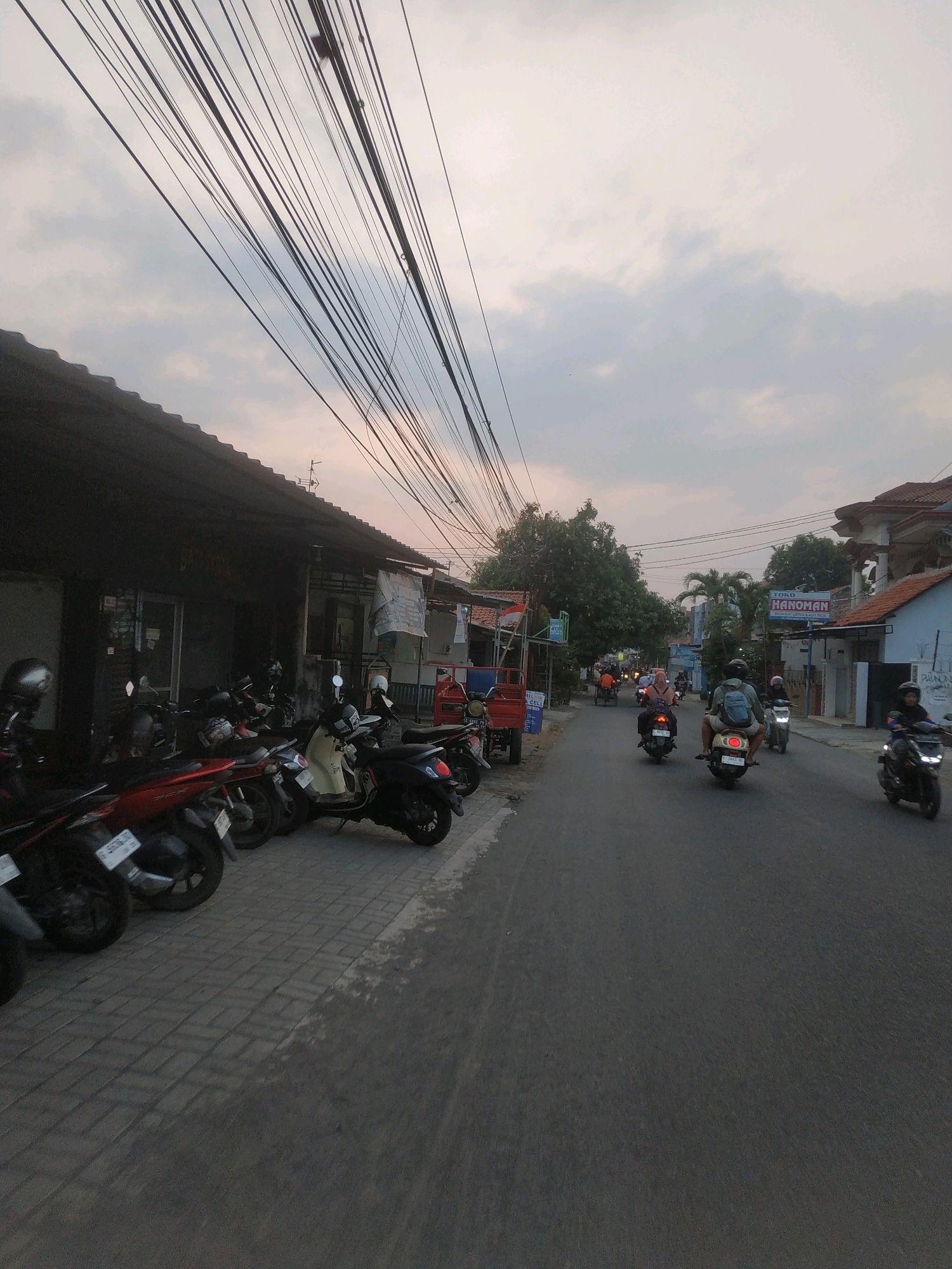 This image depicts a street scene in Tegal, Indonesia, likely during the late afternoon or early evening. The sky is overcast with a muted, yellowish hue, suggesting the setting sun. A prominent feature is the dense network of overhead electrical wires that crisscross the frame, creating a strong visual line.
On the left side of the street, a row of shops with corrugated metal roofs is visible. Parked along the sidewalk in front of these shops are numerous motorcycles, including a red scooter in the foreground, a dark-colored scooter, and several others. The sidewalk is paved with grey interlocking bricks.
The road itself is asphalt and appears to be a two-way street. Several motorcycles are in motion, with riders visible on them. In the mid-ground, a person wearing a backpack rides a scooter. Further down the road, other motorcyclists are also traveling. Buildings line the right side of the street, some appearing to be houses with tiled roofs, and one with a sign reading "TOKO HANOMAN". Green trees are scattered throughout the scene, adding to the urban landscape. A red cart is parked on the left side of the road. The overall impression is that of a bustling, everyday street in an Indonesian city.