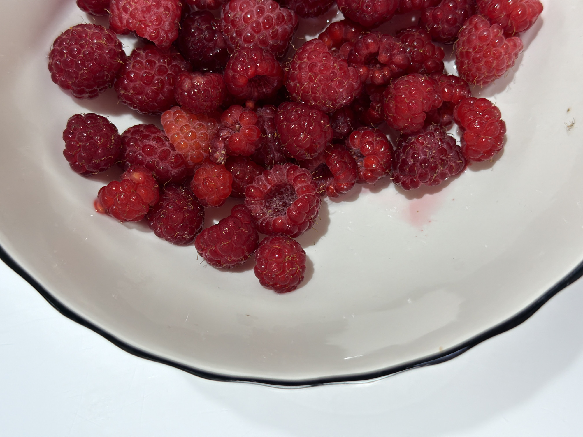 A white bowl with a black rim is filled with ripe raspberries. The bowl is placed on a white surface. The image is a close-up, top-down view of the raspberries, with the left side of the bowl visible in the foreground. The lighting is bright and casts shadows. There are no people or animals in the image. The location in the city of Chaikovsky, Russia, cannot be determined from the image.