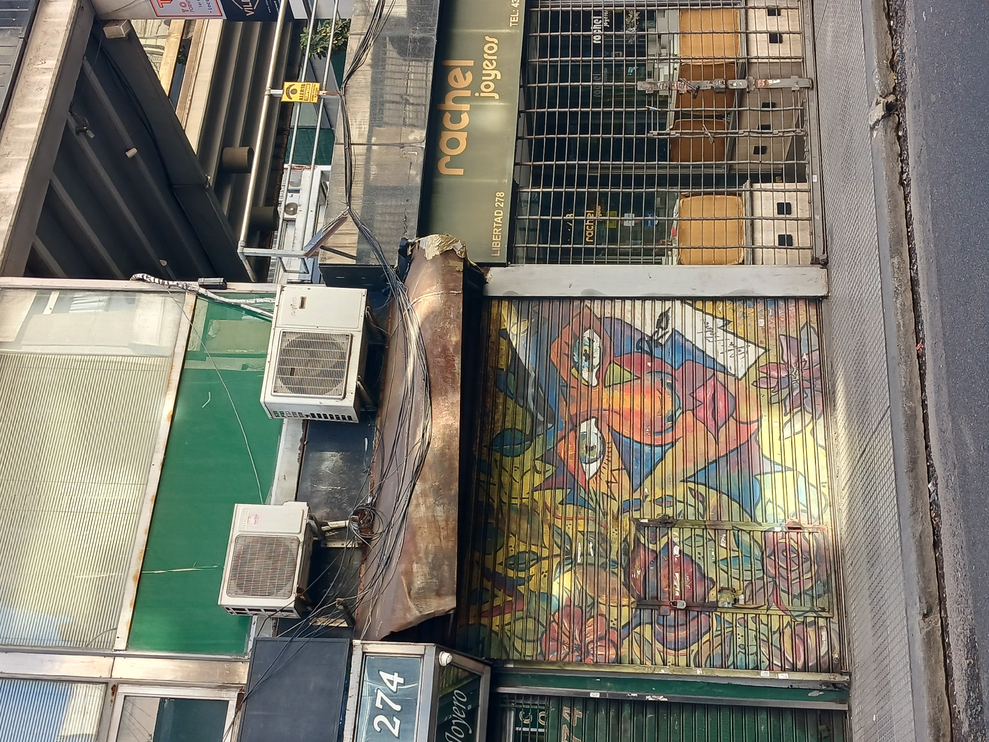 The image depicts the storefront of "rachel joyeros" located at Libertad 278 in Buenos Aires, Argentina. A metal security gate with an abstract, colorful mural covers the lower portion of the storefront. The upper portion of the shop is protected by a metal grid. Above the mural, the shop's name and address are visible on a dark green sign in gold lettering. Air conditioning units are mounted on the exterior wall to the left of the storefront, interspersed with a tangle of wires. A portion of the adjacent building at number 274 is visible on the lower left. The street and sidewalk are visible to the right.