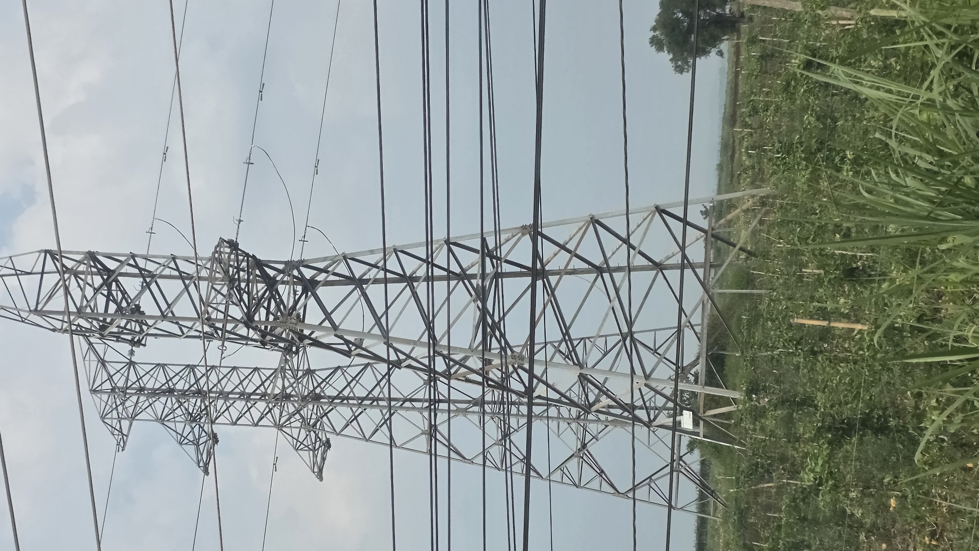 The image shows a power transmission tower situated in a rural area, likely near Xuân Long, Vietnam. The tower is made of steel and has a lattice structure with numerous cross-braces. Electrical wires are suspended from the tower. The surrounding area appears to be a field with vegetation. The sky is overcast.