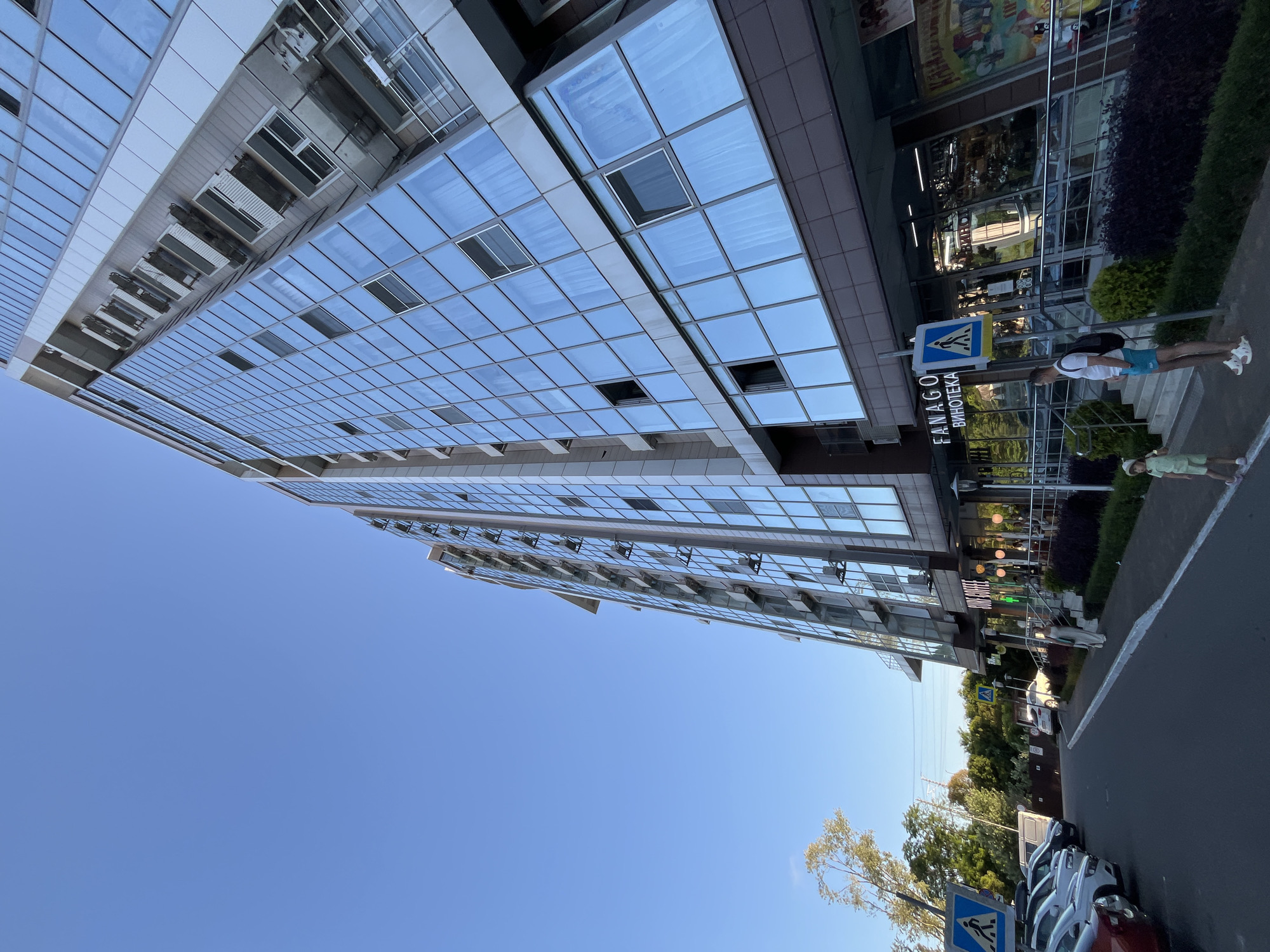 This image captures a street view in Sochi, Russia, featuring a modern, multi-story building with a glass facade and a clear blue sky. The building is flanked by a parking lot on the left, filled with several cars, and a sidewalk on the right, lined with a pedestrian crosswalk and shrubbery. The ground floor of the building houses commercial establishments, including a "FANAGO Винотека" (Fanago Wine Shop) and "SURF COFFEE". Two people are visible on the sidewalk: a man with a backpack and blue shorts, and a child in a light green outfit. The overall scene depicts an urban environment on a sunny day, likely in the afternoon given the lighting.