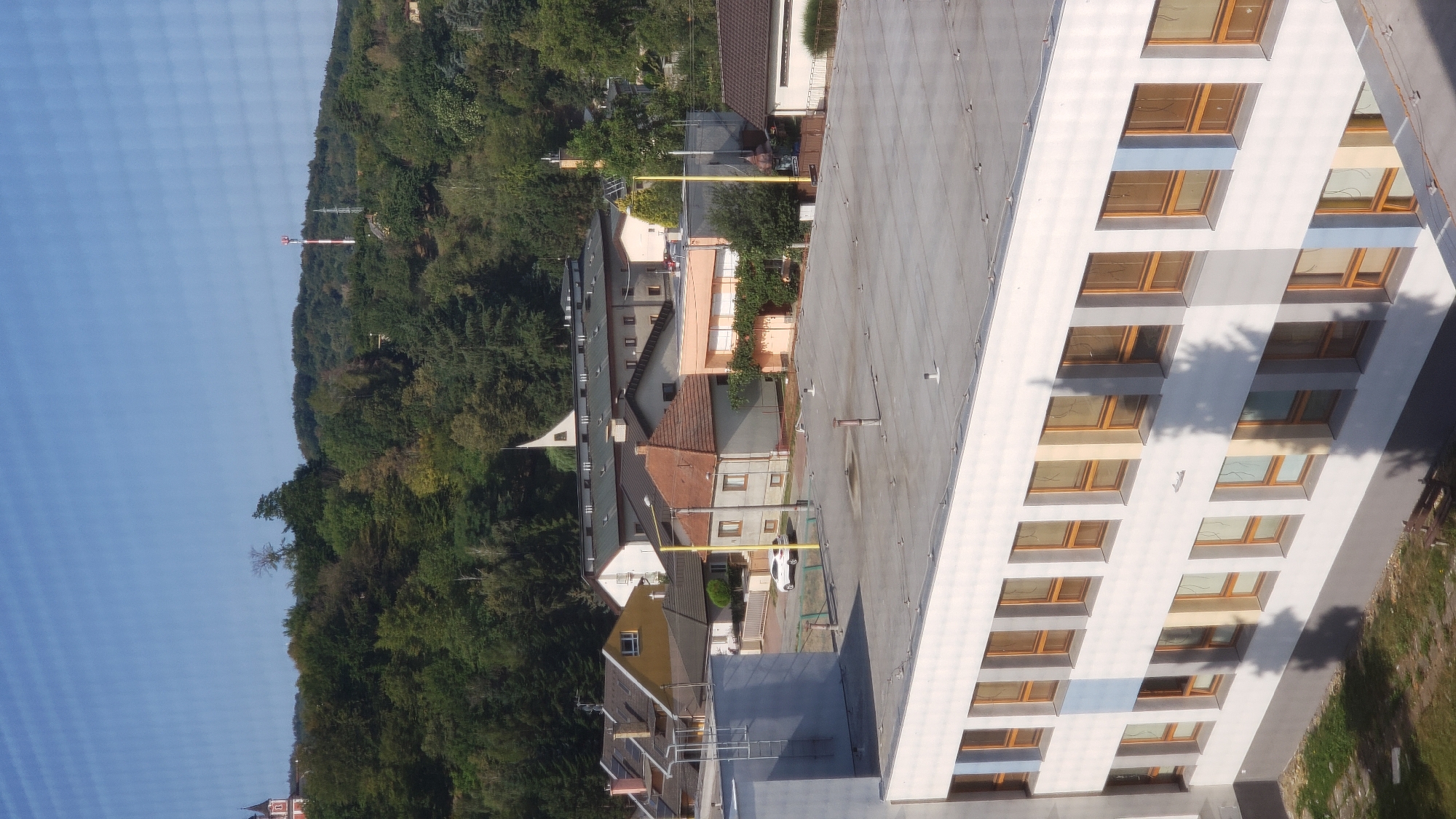 The image shows a view from a higher vantage point looking down on a town. In the foreground, a white building with multiple rows of windows is visible. The windows are rectangular with light brown frames. The building has a flat, grey roof. Beyond this building, there are several other buildings of varying sizes and architectural styles, including houses with pitched roofs. Some of these houses have solar panels installed. Further in the background, a dense green forest covers rolling hills. A communication tower with red and white sections stands on one of the hills, and a steeple of a church is visible on another. A white car is parked on a street between some of the houses. The sky is clear and blue. The scene depicts a residential or semi-industrial area in Prešov, Slovakia, characterized by a mix of modern and older buildings nestled against a natural backdrop of trees and hills.