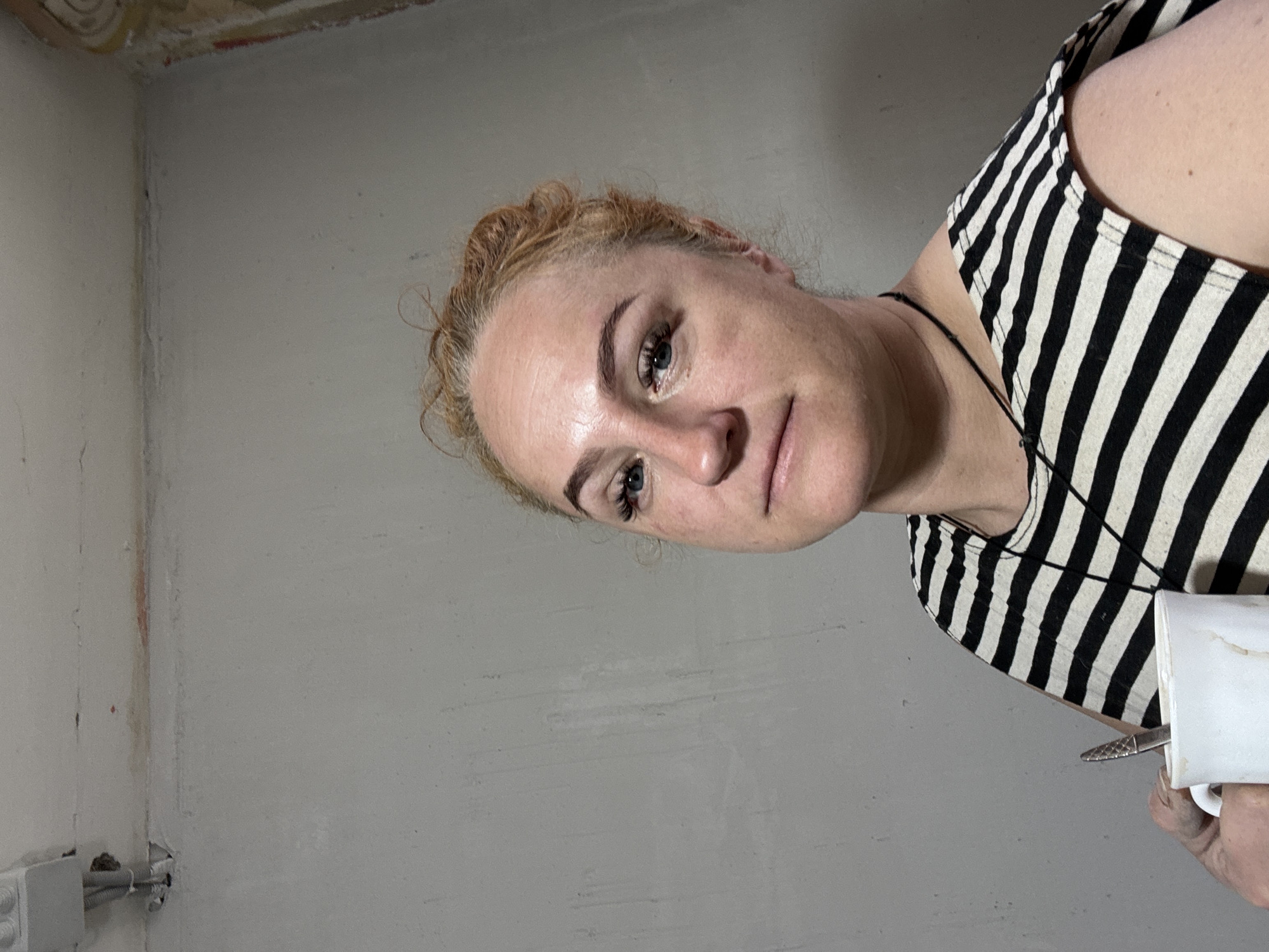 A woman with fair skin, light brown hair, and blue eyes is the primary subject, positioned at an angle in the frame. She wears a black and white striped tank top and a thin black necklace. She holds a white mug in her hand, with a metal object protruding from it.
The background consists of two intersecting walls, painted in a muted gray color. The upper corner of the walls shows unfinished sections with exposed construction materials. Near the bottom left corner, there are electrical conduits and a white junction box attached to the wall. The location is possibly a room undergoing renovation in the city of Chaikovsky, Russia.