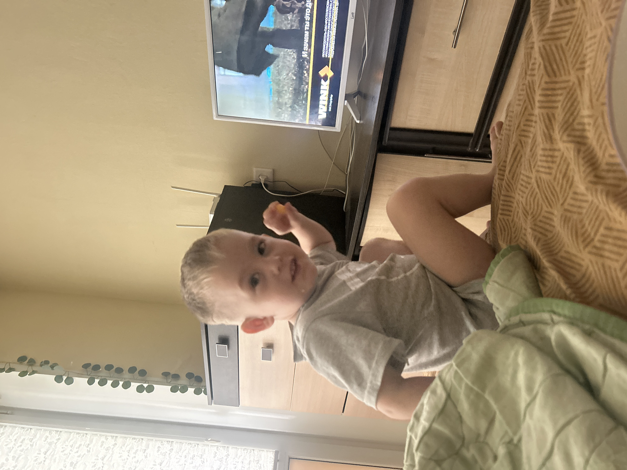 An indoor scene depicts a young child in a domestic setting. No specific visual cues within the image definitively confirm the location as Chaikovsky, Russia; however, the television display includes text in Russian, featuring the "WINK" streaming service logo and a reference to "PAO 'Rostelecom'," indicating a Russian context.
In the foreground, a light green quilted blanket is partially visible on the left. A fair-skinned toddler with short, light-colored hair, wearing a light grey t-shirt and light-colored shorts/underwear, sits on a brown blanket with a repeating geometric line pattern. The child's body is angled slightly to the right, but their head is turned towards the camera with a neutral expression. A small, round, yellowish-orange object is held in the child's right hand.
In the midground, to the left of the child, a chest of drawers is visible. Its top drawer is dark grey with a square silver handle, and the two drawers beneath it are light wood-toned with identical grey square handles. To the right, a dark, flat surface supports a black electronic device (likely a Wi-Fi router) with two white antennas, connected to a white wall outlet via a white cable. Adjacent to this, a white flat-screen television, branded "Leff," is on. The television screen displays an image with Russian text, including "WINK" and "И зачем ты это дела?" (And why did you do it?), along with an advertiser's notice for "PAO 'Rostelecom'."
In the background, a light yellowish-beige wall is visible. To the far left, a window with white blinds is partially obscured by the frame, and a decorative string of artificial green leaves hangs vertically along the wall next to it.