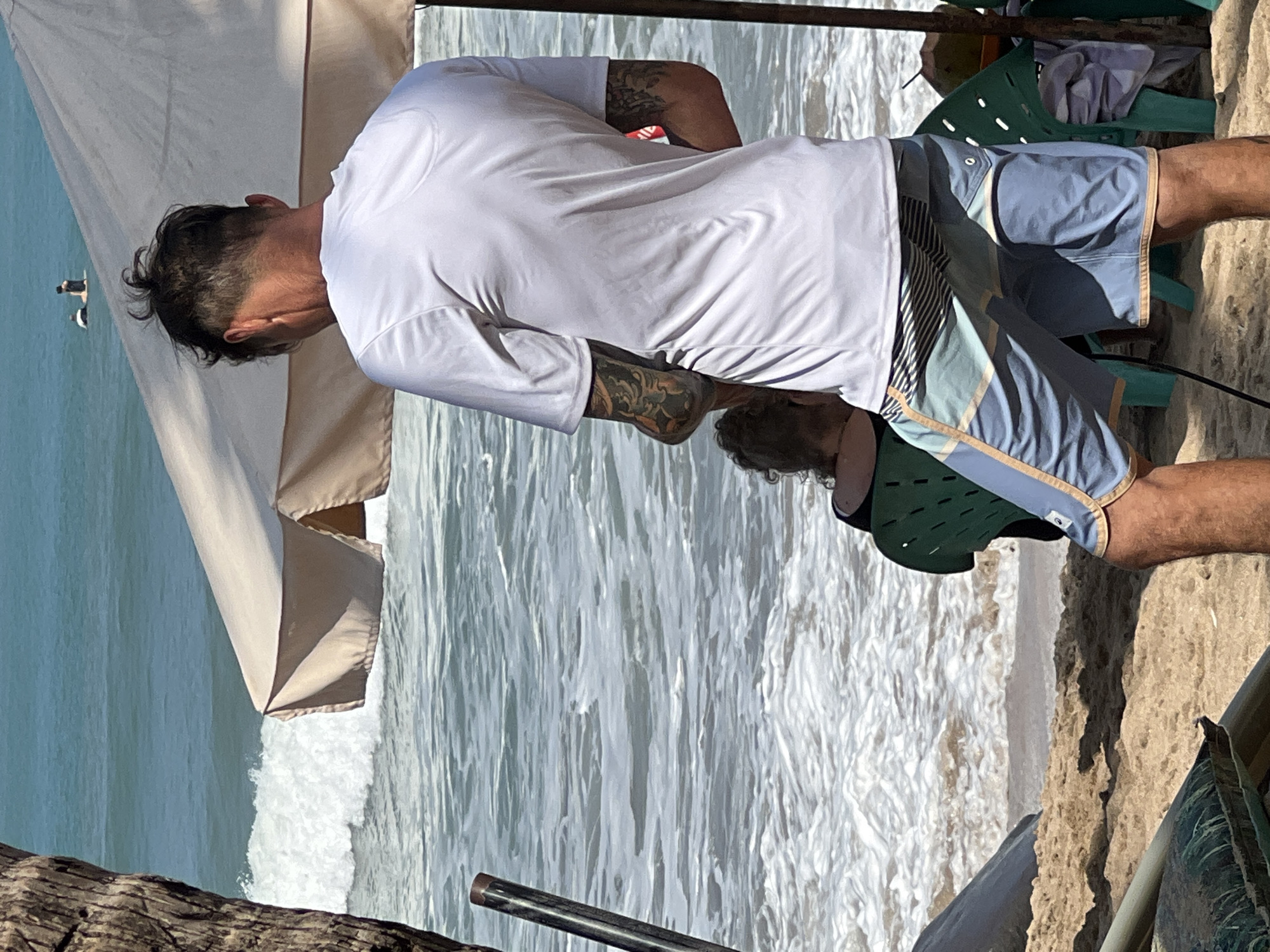 In Kuta, Indonesia, on a sunny day, a man with tattoos on his arms and a messy bun hairstyle stands on a sandy beach facing the ocean. He is wearing a white t-shirt and blue swim shorts with a patterned waistband. In front of him, a person with curly hair is seated in a green plastic chair, facing away from the camera. A large beige umbrella provides shade over the scene. The ocean waves are crashing on the shore, and two surfers can be seen in the distance. The overall environment suggests a casual beach setting with the ocean as the prominent backdrop.
