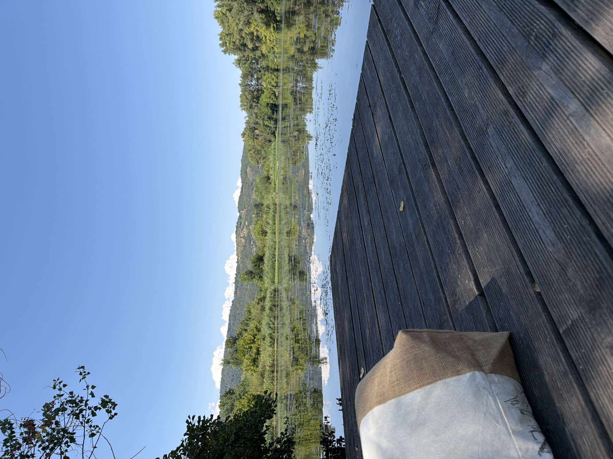 An outdoor, low-angle shot depicts a wooden dock in the foreground. A beige and white cushioned object sits on the left side of the dock. The dock extends towards a calm body of water filled with reflections of the sky and surrounding greenery. Trees and hills form the background under a clear blue sky with scattered white clouds. The location is the River Rhône at Tournon-sur-Rhône, France.
