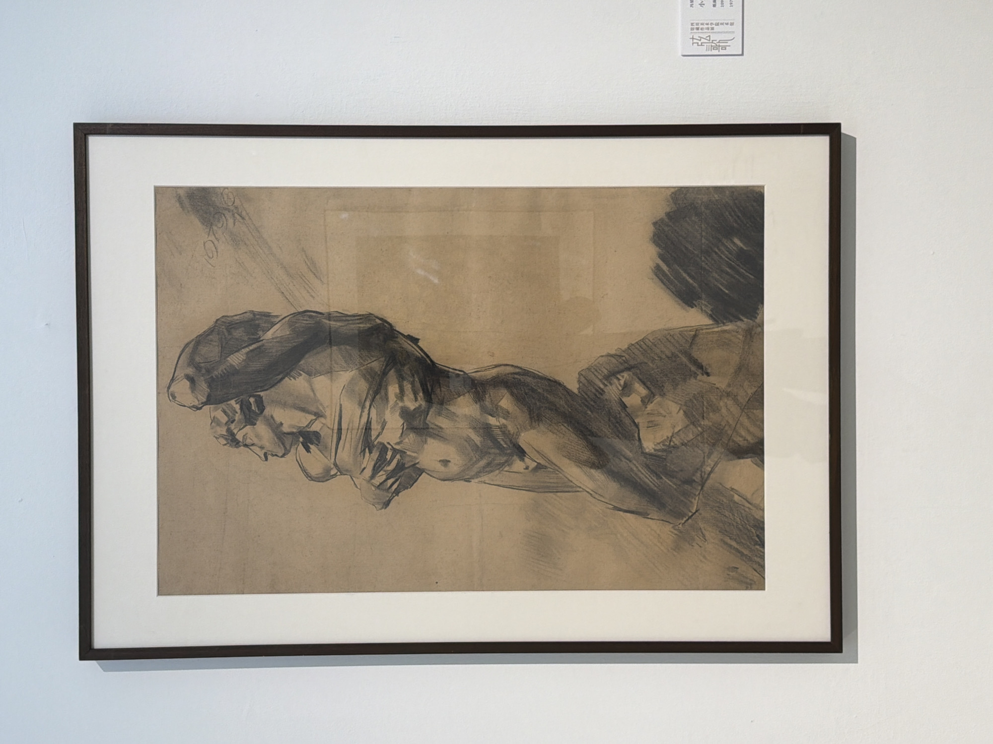 A framed charcoal drawing of a male figure is displayed on a white wall. The drawing depicts a muscular man in a dynamic pose, with his arms crossed over his chest, looking upwards. The figure is rendered in shades of grey and black on a tan paper background, with visible charcoal strokes and smudging. The man appears to be standing on a rocky or irregular surface, which is also depicted with heavy charcoal shading. The drawing is matted with a thick white border and framed with a dark, narrow frame. To the right of the framed artwork, a small white label with text is affixed to the wall. The label contains Chinese characters and numerical information, likely an artwork description or information. The lighting appears to be artificial, casting a soft illumination on the artwork and the wall. The context suggests this is an exhibition or gallery setting.