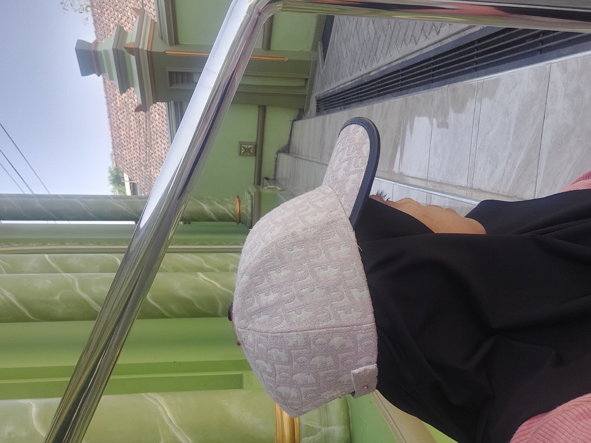 A person wearing a black hijab and a light pink cap with a repeating pattern is partially visible in the foreground, looking to the right. The cap has a dark brim. The person is positioned next to a light green wall with decorative vertical elements and a silver railing in front. In the background, there is a building with a tiled roof, a prominent decorative pillar, and more light green walls. The environment appears to be outdoors, possibly a courtyard or an area next to a building in Sedan, Indonesia, given the architectural style and the presence of a mosque-like structure. The ground is paved with light-colored tiles. The lighting suggests a sunny day.