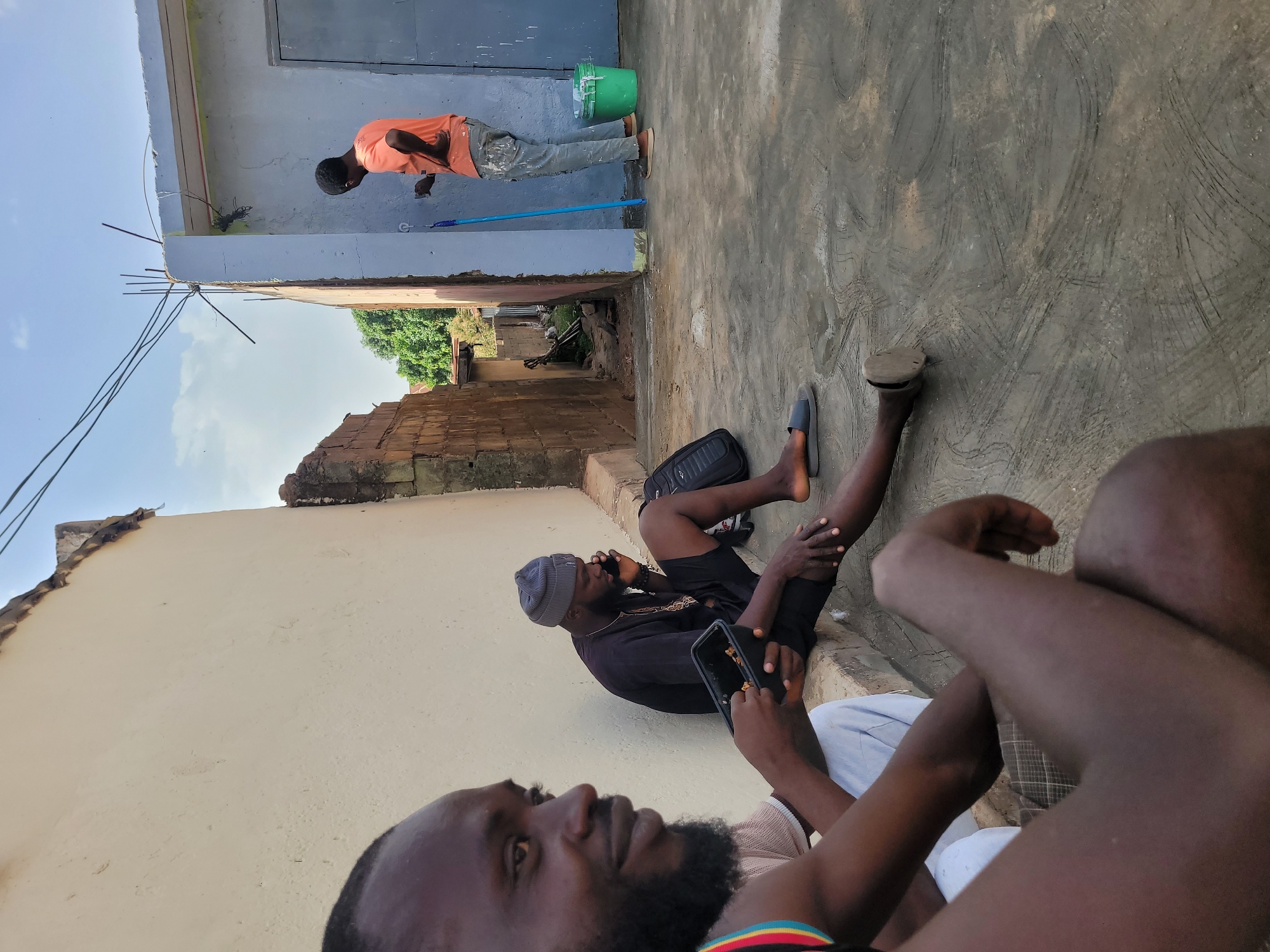 This media file captures an outdoor scene, likely in Jos, Nigeria, showing three men engaged in different activities on a bright, sunny day with a clear blue sky.
In the foreground, the profile of a man's face, neck, and shoulder is visible on the right, looking slightly to his left. He has a dark beard and wears a shirt with a striped collar. Part of another person's leg and hand are also visible close to him.
In the mid-ground, another man is seated on the ground or a low ledge, facing generally towards the right. He wears a black shirt, dark shorts, and a grey beanie. He is holding a mobile phone to his left ear while another phone rests in his right hand or lap. A black backpack lies beside him, and his bare feet, one next to a dark slipper, are visible.
In the background, positioned to the right, a third man stands, appearing to be working on a light blue-grey wall. He wears an orange t-shirt, paint-splattered grey pants, and light-colored shoes. He holds a long blue pole-like tool, possibly for painting or cleaning. A green bucket sits on the ground next to him.
The setting features concrete or cement ground surfaces with textured patterns. Various building structures are visible, including plastered walls and sections of exposed brick. An alleyway or path recedes into the distance, bordered by walls, revealing trees and distant structures. Unfinished construction elements, such as rebar protruding from a concrete slab, are present on the left side of the image. The overall atmosphere is relaxed, with one person working while others rest. No legible text is visible.
