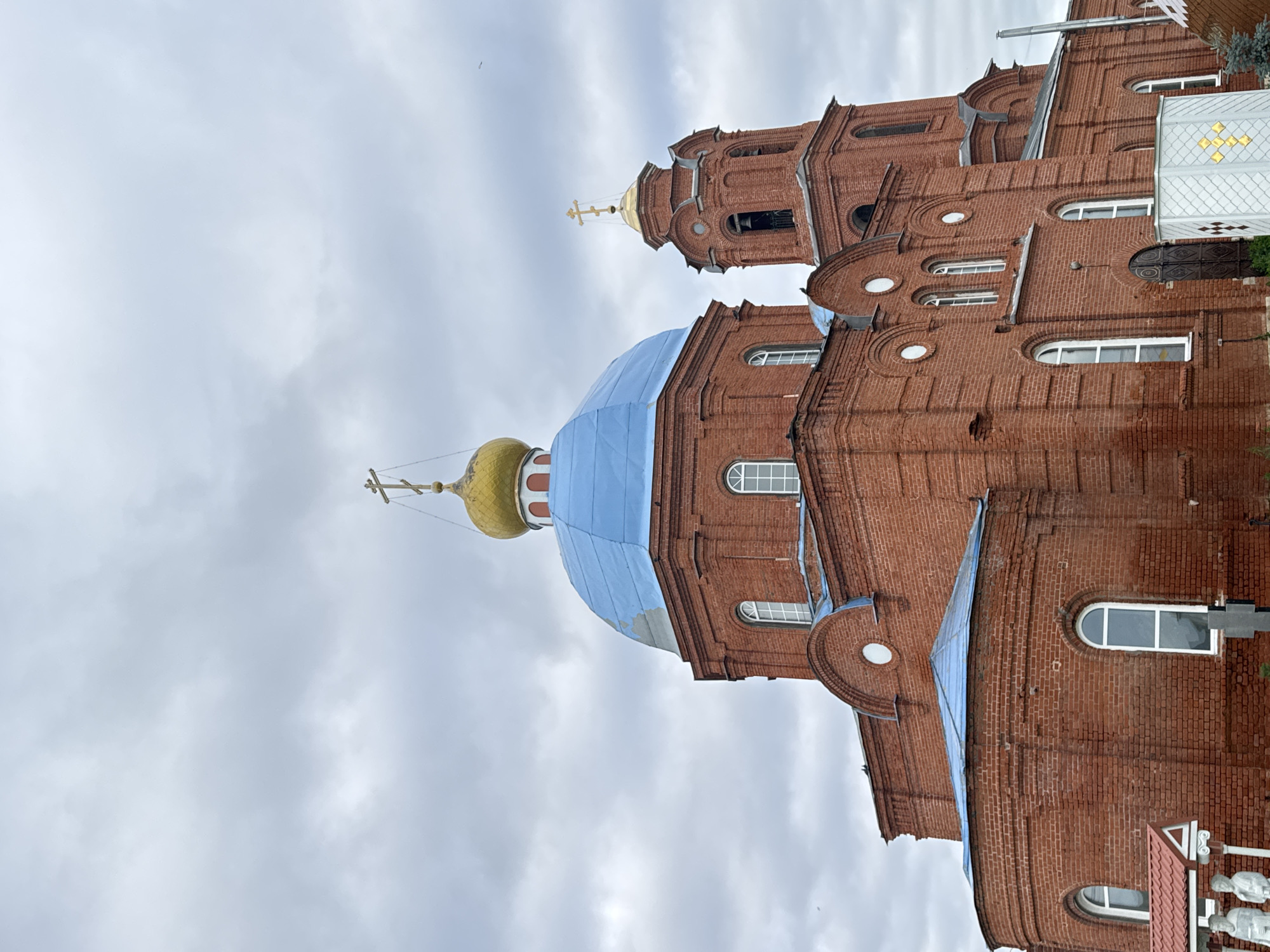 The image shows a brick church in Anateh, Russia. The church has a blue dome and a golden cross on top. There is also a bell tower with a golden cross on top. The sky is cloudy.