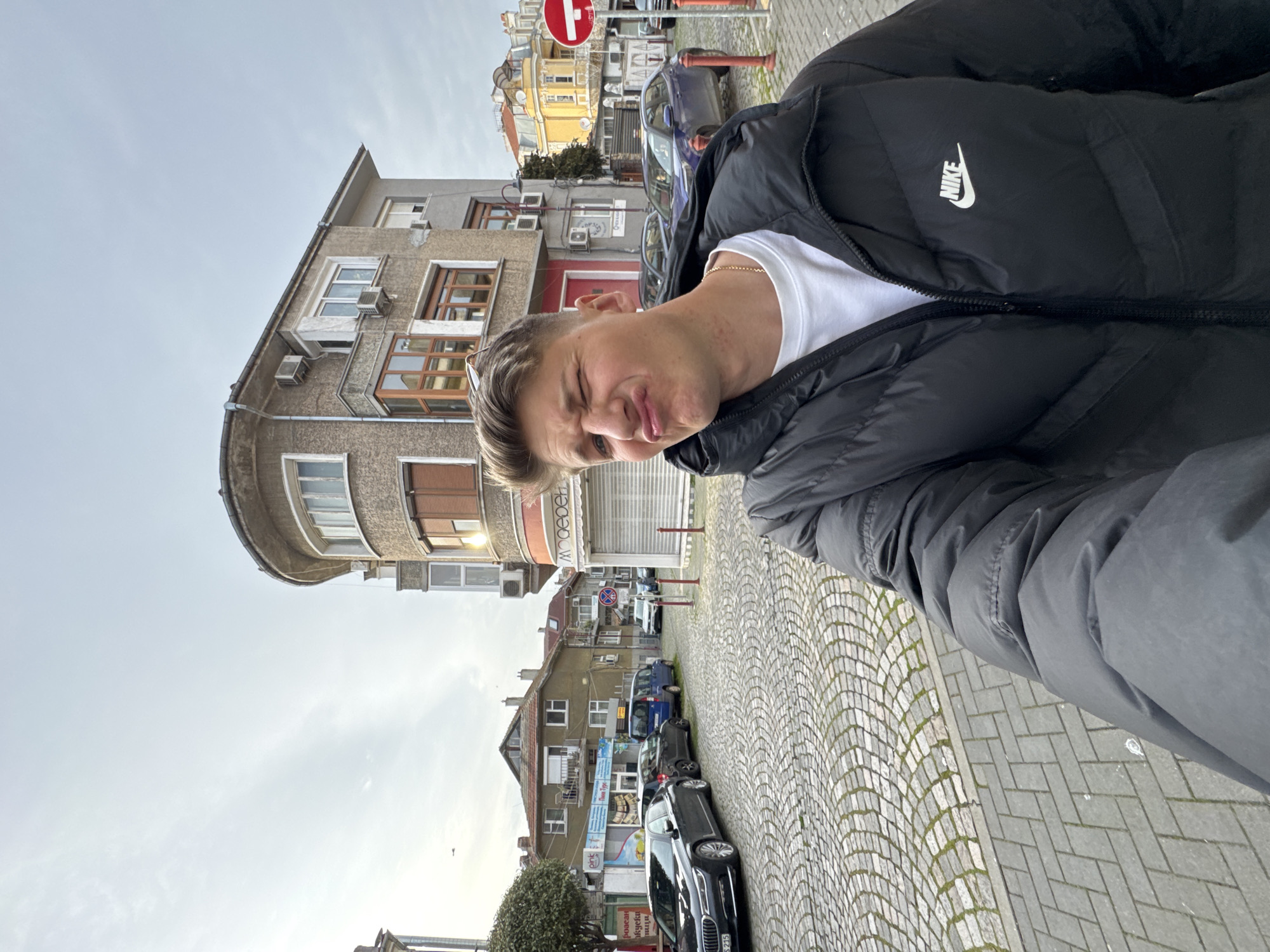 A young man stands on a cobblestone street in Burgas, Bulgaria. He wears a black Nike puffer jacket and a white t-shirt. A gold chain is visible around his neck. The man is making a comical face, with his eyes squinted and lips pursed. Behind him, older buildings line the street. Cars are parked along the curb. There is a no entry sign on the right side. Shops with closed metal shutters are visible. The sky is overcast.
